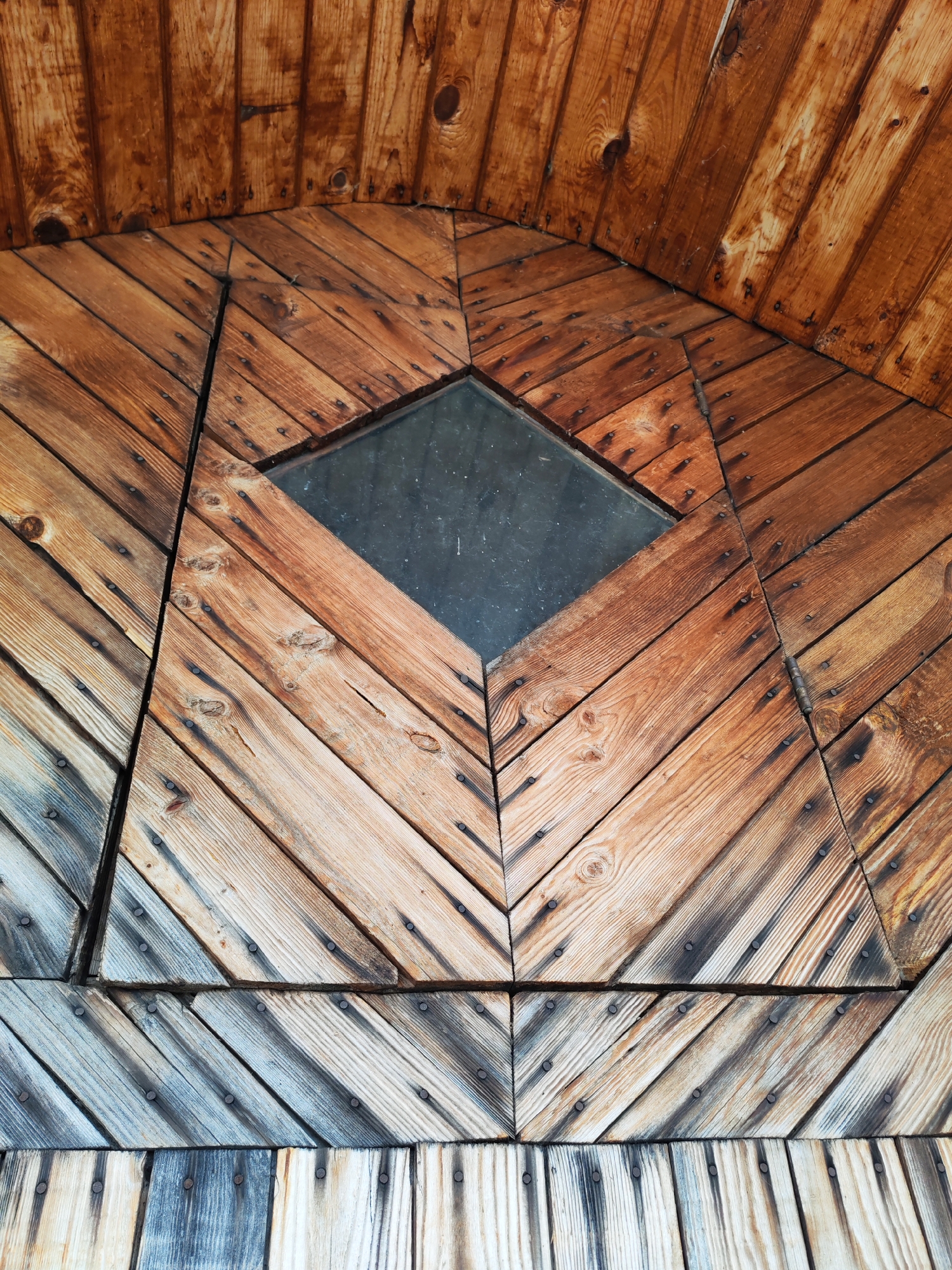 The image shows a detailed view of a wooden structure, possibly a door or architectural element, constructed with diagonally arranged wooden planks. A diamond-shaped glass insert is centrally located. The wood varies in tone from light to dark brown with some areas showing graying and weathering, particularly towards the bottom. The planks are affixed with numerous nails, which are visible throughout the structure. The arrangement is symmetrical, forming an arch-like pattern. The location is specified as Chaikovsky, Russia, but this cannot be confirmed solely from the visual content.