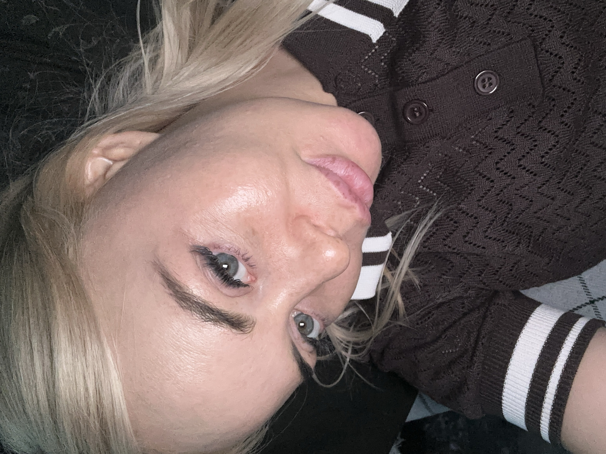 An adult female with blonde hair and fair skin is depicted in a close-up, tilted self-portrait, facing the camera. Her eyes are light greenish-blue with visible eyelashes and dark, defined eyebrows. Her lips are full and naturally pink, slightly parted. The skin on her face exhibits visible texture and a slight sheen, particularly on the forehead and left cheek. She is wearing a dark brown, short-sleeved, open-knit top featuring a zigzag or chevron pattern throughout the fabric. The top has a polo-style collar with a two-button placket, showing two dark buttons. The sleeves have ribbed cuffs with two prominent white horizontal stripes. The background is dark and indistinct, suggesting an indoor setting, possibly with a dark fabric or bedding. A small portion of a grey and white plaid-patterned fabric is visible in the lower left corner. The lighting appears to be frontal, illuminating her face. The location is in the city of Unknown, Unknown.