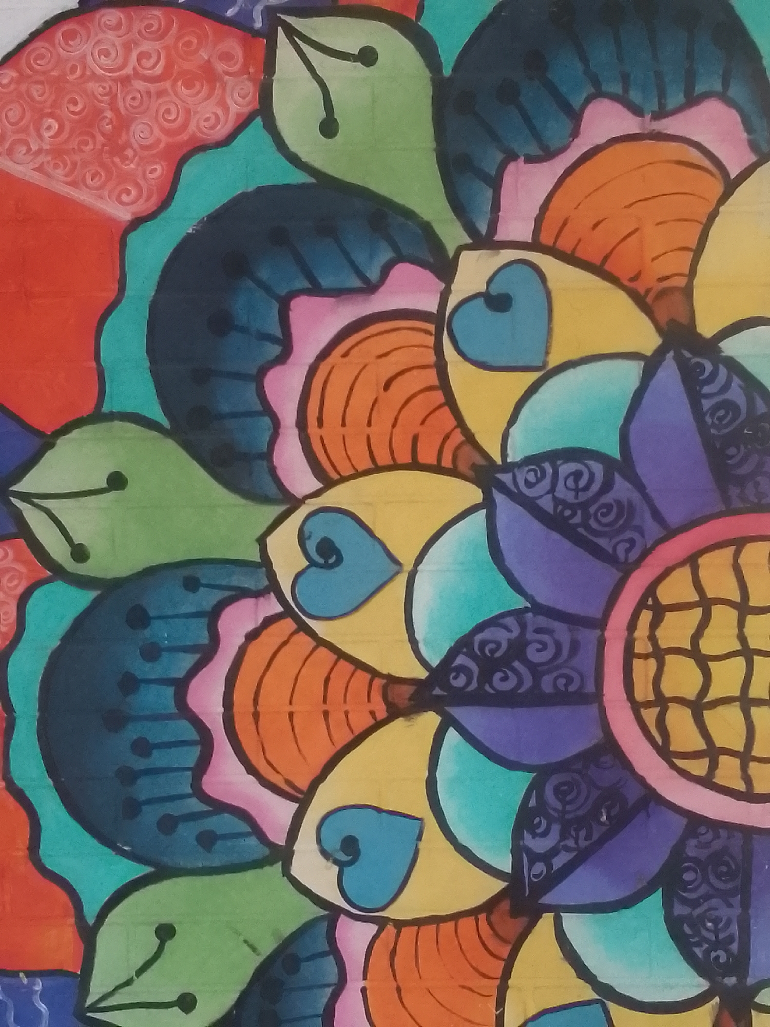This close-up image captures a vibrant, colorful, stylized artwork resembling a floral or mandala pattern. The composition is formed by numerous interlocking and overlapping petal-like segments, all distinctly outlined with thick black lines.
A rich and diverse color palette is utilized, featuring bright reds, oranges, yellows, various shades of green (including lime and olive), blues (teal, dark blue, and light blue), pink, and purple. Several segments showcase intricate details: the red area on the far left has white swirling spiral patterns; certain dark blue sections exhibit parallel lines and small dots; green leaf-like shapes contain simple veins and dots; yellow petals feature light blue heart shapes; and purple segments are adorned with darker purple spiral motifs. Towards the right-center, a circular element with a yellow and orange grid or waffle pattern is visible, encircled by a pink border.
The artwork appears to be created on a textured surface, possibly paper, as faint grid lines are perceptible beneath the colors, especially in the lighter-toned areas. The overall design emphasizes radial symmetry, giving it the appearance of a blossoming flower or an abstract mandala. No people, animals, or visible text are present in the image. The scene is static, focusing solely on the intricate artistic design. The cropped nature of the image suggests it is a detail from a larger artwork. The artwork is located in Juan Griego, Venezuela.