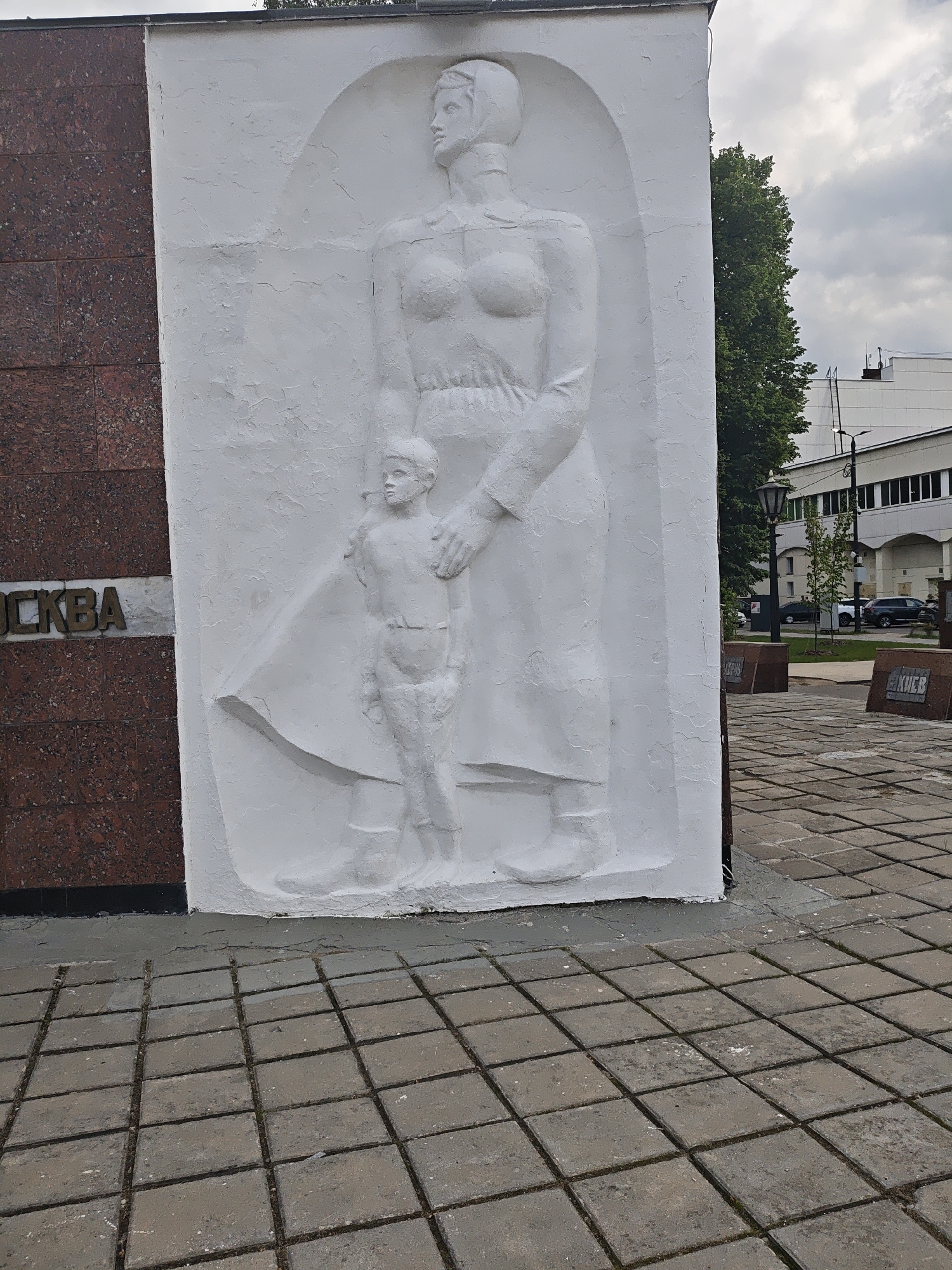 A low-angle, medium shot features a portion of a monument in Noginsk, Russia. On the left is a segment of red granite, with the word "OCKBA" in tarnished gold-colored letters. Adjacent is a white concrete relief depicting a mother with a child. The woman has a headscarf and a dress or robe, and is standing. She has one arm holding the child. The child is small, standing beside the mother. The base of the monument is set on a paved surface composed of grey square blocks. Behind the monument, there's a street with parked cars and buildings in the background, with a few trees visible. The sky is overcast. Additional markers are on the pavement.