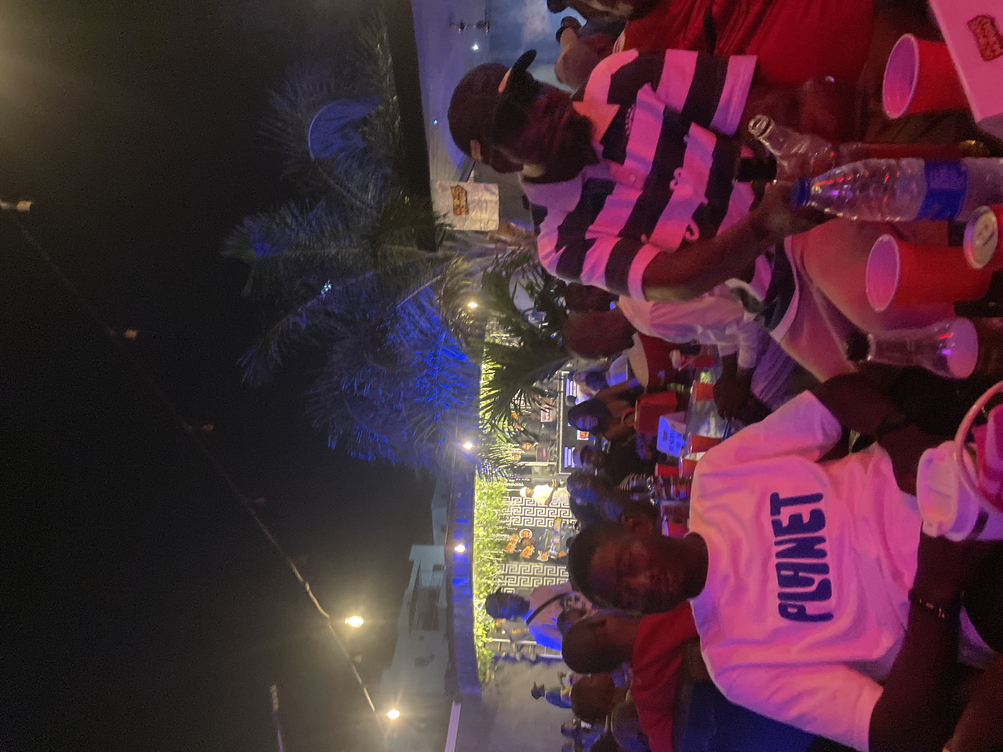 The image depicts a nighttime gathering at an outdoor venue in Guzape, Nigeria. The scene is illuminated by string lights and vibrant colored lighting, casting a warm and festive atmosphere. In the foreground, a man wearing a white t-shirt with the word "PLANET" printed in blue is seated, facing the camera. To his right, another man in a black baseball cap and a horizontally striped shirt is also visible. The background reveals a stage area where a band is performing, with people gathered around tables and mingling. Palm trees and architectural elements suggest an urban setting. Red plastic cups are prominent on the tables, along with bottles of water and other beverages. The overall impression is of a lively social event or concert taking place after dark.