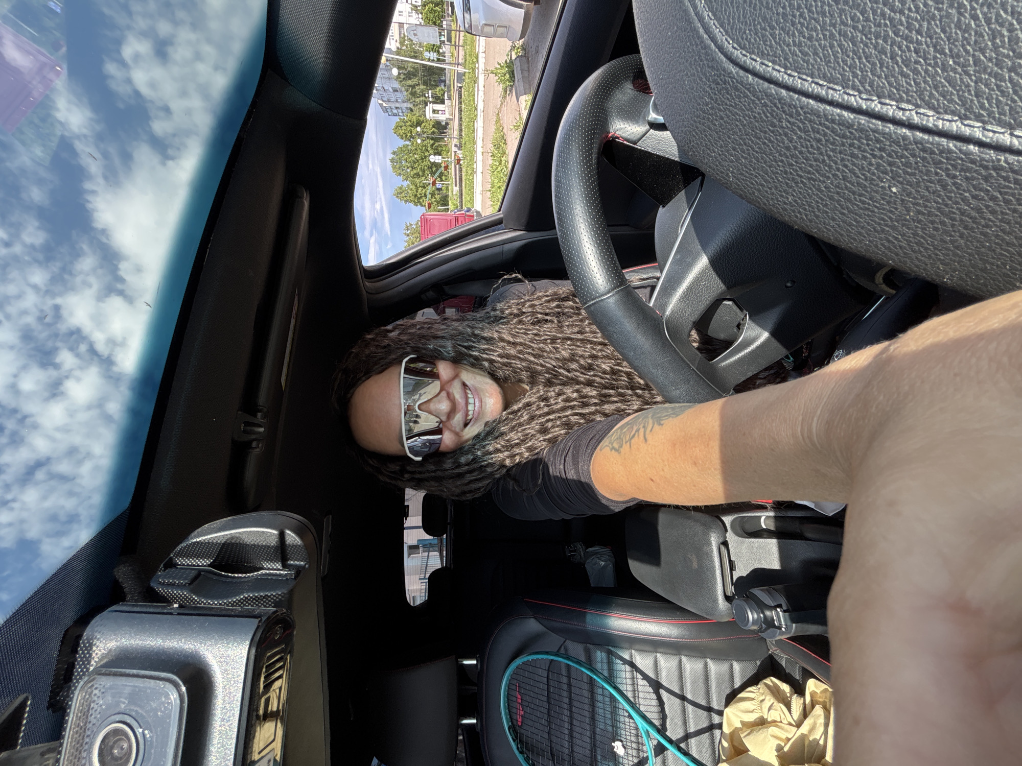 The image shows an interior car scene, taken from the driver's seat with the camera pointed towards the passenger side and partially upwards. A person with long dreadlocks and sunglasses is visible in the passenger seat. A steering wheel is visible in the top right of the image.
In the foreground, the dashboard with a camera is visible, with a tennis racket and a crinkled paper bag on the passenger seat. Through the car window, buildings and trees can be seen, indicative of an urban environment. Based on these details, it can be hypothesized that the location may be the city of Chaikovsky, Russia.