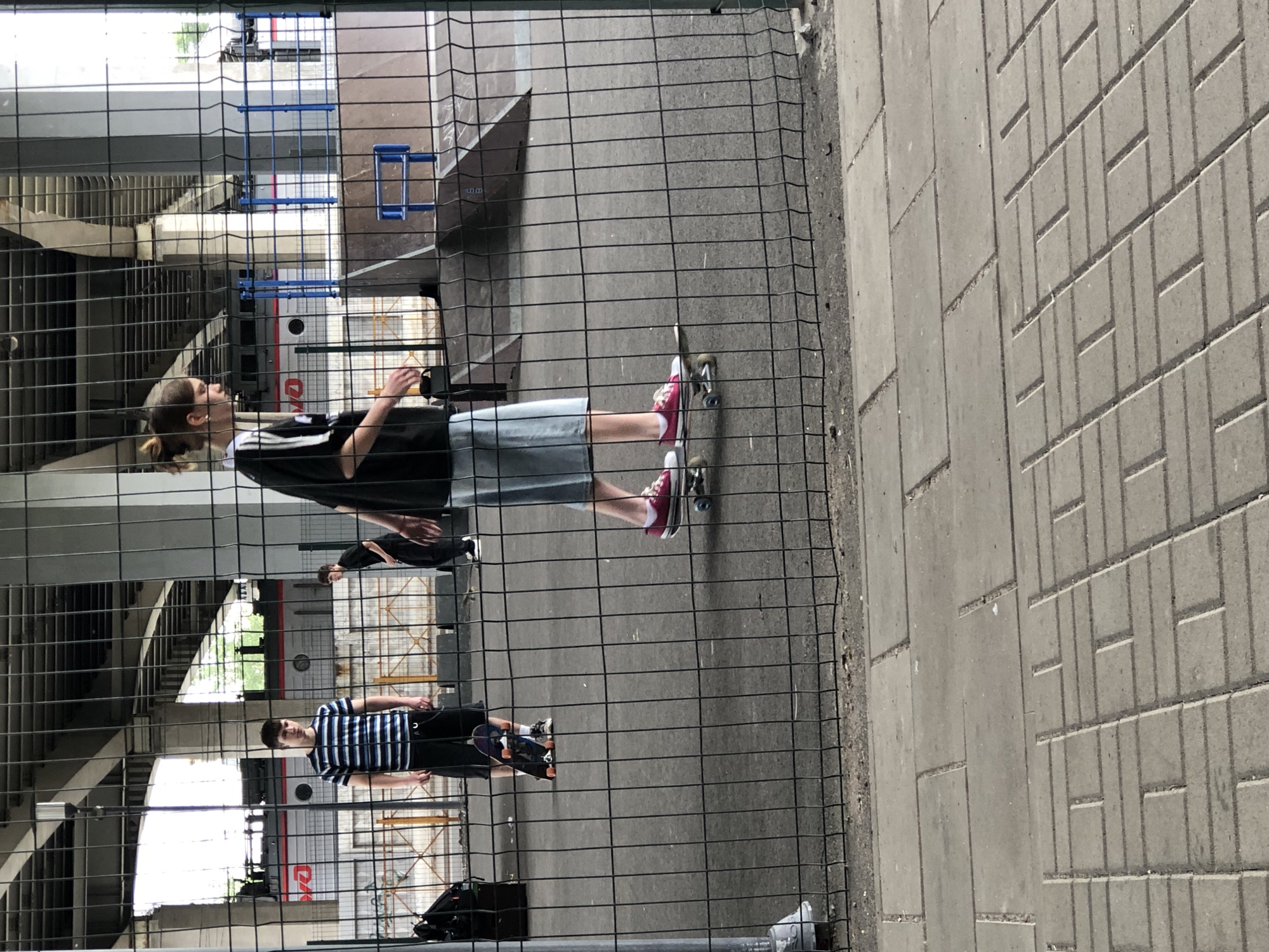 The image shows a skateboarding park located under an overpass in Saint Petersburg, Russia. A young person is skateboarding in the foreground, viewed from the side. They are wearing a black t-shirt with white stripes on the sleeves, light blue denim shorts, and pink sneakers. Another person, a young male, is standing to the left, facing forward, wearing a striped t-shirt and dark shorts. In the background, another young male is visible, wearing dark clothing. The park has ramps and a metal fence surrounds the area. A train is partially visible in the background, with the Russian Railways logo on its side. The ground is paved with interlocking grey concrete blocks.