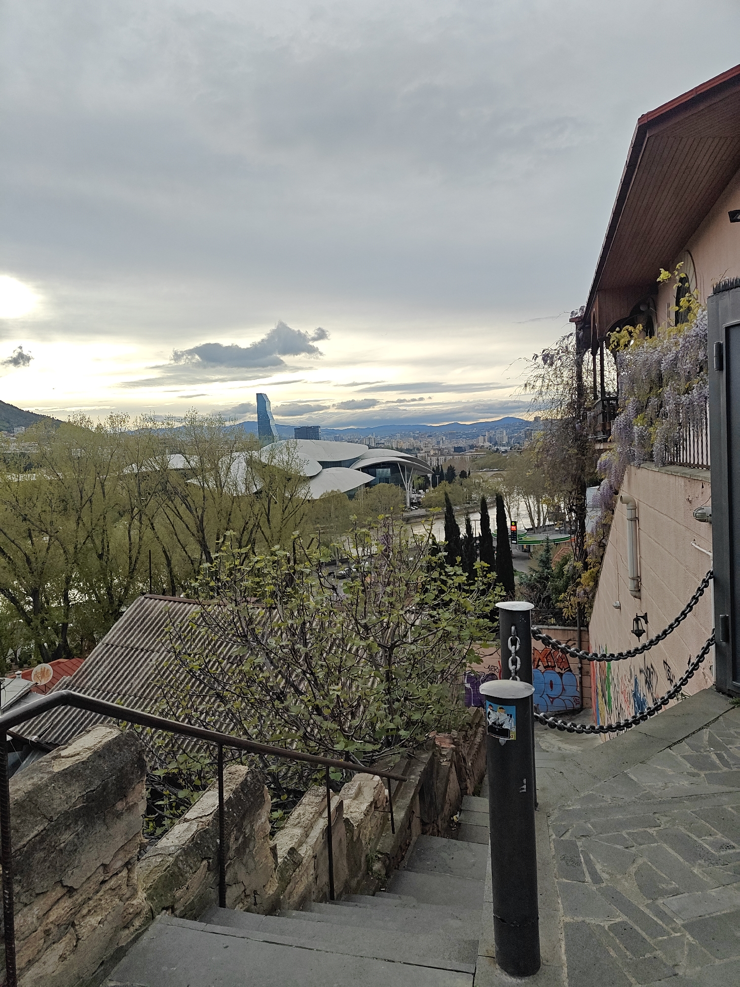 This photograph captures an elevated view overlooking a vibrant urban landscape in Tbilisi, Georgia, under an overcast sky. In the immediate foreground on the left, stone steps with a metal railing descend, bordered by a rough stone wall. To the right, a multi-story building with a light pink facade and a brown-tiled roof features intricate wooden balconies and is heavily adorned with blooming purple wisteria. A black metal pole stands in the foreground, connected by a heavy black chain to the building's wall, below which colorful graffiti covers the lower section of the wall. One visible piece of graffiti reads "LOVER". A small sticker with cartoon figures is adhered to the pole.
The midground is dominated by lush green trees, likely with new spring foliage, that partially obscure a river flowing through the city. Beyond the trees and river, several distinctive modern buildings are visible, including the wave-like structures of what appears to be the Public Service Hall (Palace of Justice) and a tall, slender glass skyscraper. A gas station with a visible sign can be seen near the riverbank. Rolling hills and mountains form the distant backdrop, blending into the hazy horizon. The sky is largely covered with thick, grey clouds, though brighter areas on the left suggest the sun is either low in the sky or attempting to break through the cloud cover, indicating late afternoon or early evening. The overall scene presents a blend of old-world charm, natural elements, and contemporary urban development.