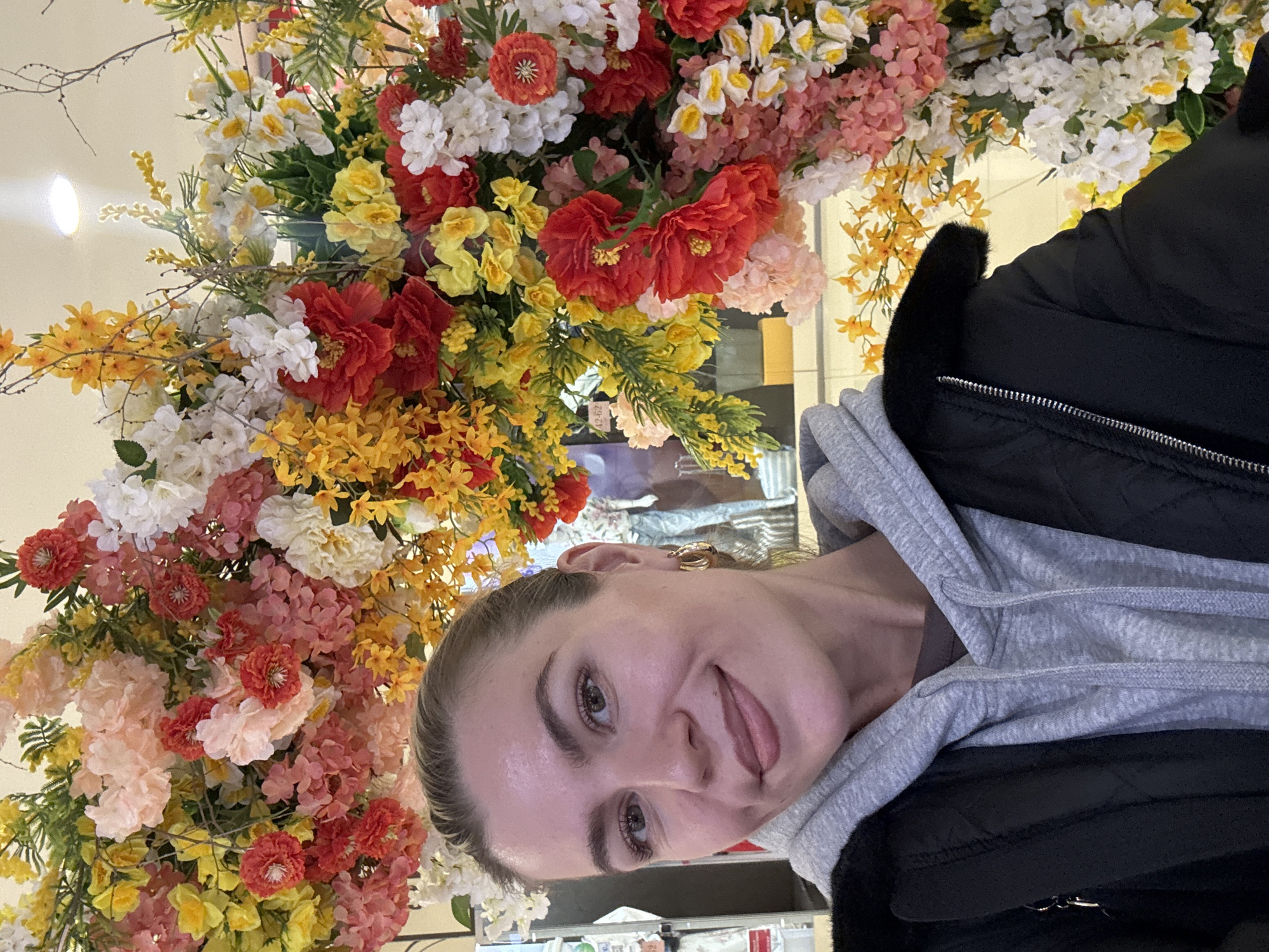 The image is a selfie of a woman with a large arrangement of artificial flowers in the background. The woman has light skin, brown eyes, and her hair is pulled back. She is wearing a gray hooded sweatshirt under a black jacket with a furry collar. Her tongue is slightly out.
The floral arrangement behind her is vibrant and colorful, featuring a mix of red, yellow, orange, white, and pink flowers with green foliage.
The location appears to be indoors, likely in a shop or commercial space, but the specific location in the city of Unknown, Unknown is not discernible from the image. There is a reflective surface behind her, which could be a window or display.
