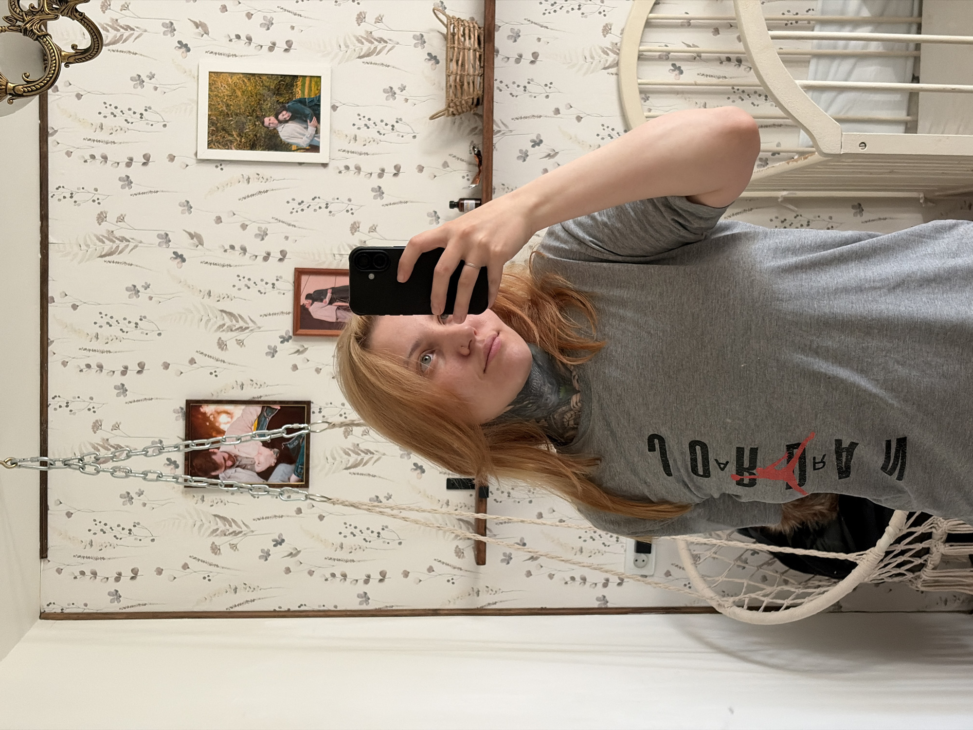 A woman is taking a selfie in a room with floral wallpaper. The wallpaper features small botanical patterns in muted colors. Several framed photos hang on the wall behind her, along with a wall-mounted basket. She is wearing a gray t-shirt with reversed lettering, and a neck tattoo is visible. She is holding a black smartphone in her right hand. A white crib is partially visible in the upper right corner, and a white hammock chair hangs in the lower right corner. An electrical outlet is visible near the bottom right of the image. Based on the location metadata, this photo was taken in the city of Chaikovsky, Russia.