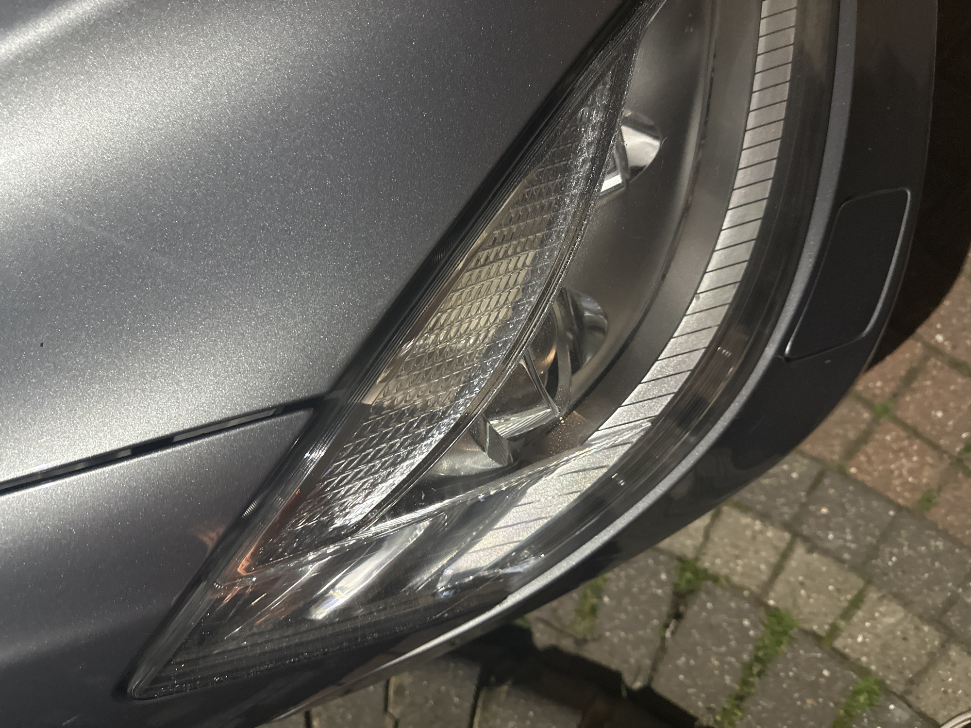 The image presents a close-up, downward-angled view of the front driver's side of a metallic grey passenger vehicle, likely a car, under artificial low-light conditions. The prominent subject is the vehicle's complex headlight assembly, which features clear lenses revealing internal projector elements, a textured or diamond-patterned upper section, and a lower integrated strip with horizontal opaque lines. The adjacent metallic grey paintwork of the fender and bonnet shows a fine sparkle. A segment of the matte black plastic bumper is visible directly below the headlight, incorporating a rectangular recessed panel, possibly for a headlight washer or sensor. The vehicle is positioned on an outdoor ground surface composed of interlocking concrete block pavers, displaying varied shades of grey and reddish-brown. Sparse green vegetation, consistent with weeds or grass, is visible growing in the gaps between the pavers. No individuals, specific actions, or distinct geographical markers are present in the image to confirm the location as the city of London, United Kingdom, though the paving type is common in urban environments.