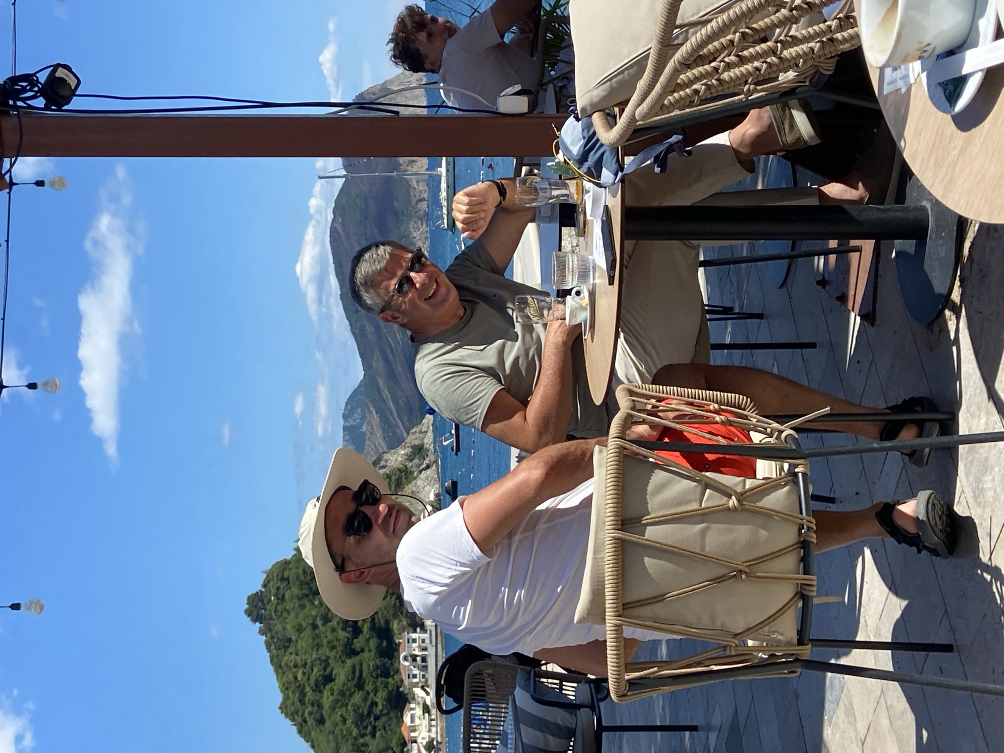 In Petrovac centar, Montenegro, two men are seated at a table outdoors, facing the sea. The man on the left wears a white t-shirt and a wide-brimmed hat with sunglasses. The man on the right, also wearing sunglasses, is dressed in a grey t-shirt and light-colored shorts. He is smiling and appears to be talking, gesturing with his right hand. On the table between them are glasses, a cup, a phone, and what appears to be a drink. A third man is visible in the background, seated at another table. The scene is set against a backdrop of the Adriatic Sea, with boats and distant hills. The area is a resort or cafe with outdoor seating and overhead string lights.