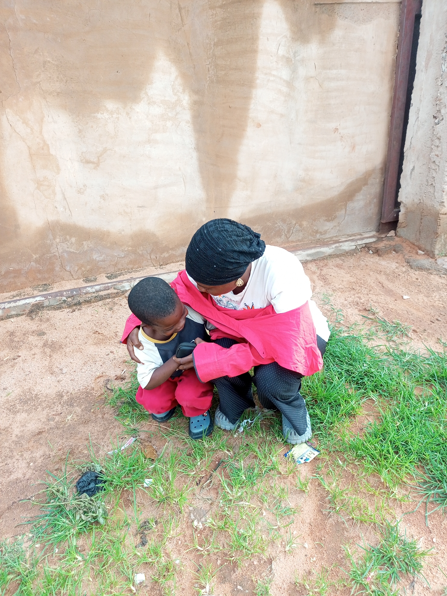 A person wearing a black head covering, a white t-shirt, and a pink jacket is crouching down, embracing a young child who is also crouching. The child wears a t-shirt with white, black, and yellow sections, red pants, and grey shoes. The person holding the child is wearing black pants with a white polka dot pattern and grey shoes. Both are looking down at something the child is holding in their hands, possibly a smartphone. The background is a rough, beige wall with some staining and a metal door frame on the right. The ground in front of them is sandy dirt with patches of green grass, and there appears to be some litter on the ground. The scene suggests a candid moment, with the person appearing nurturing towards the child. The lighting appears to be natural daylight, suggesting it is daytime. The location cues, such as the wall texture and the type of vegetation, are consistent with an outdoor setting in Tempure, Nigeria.