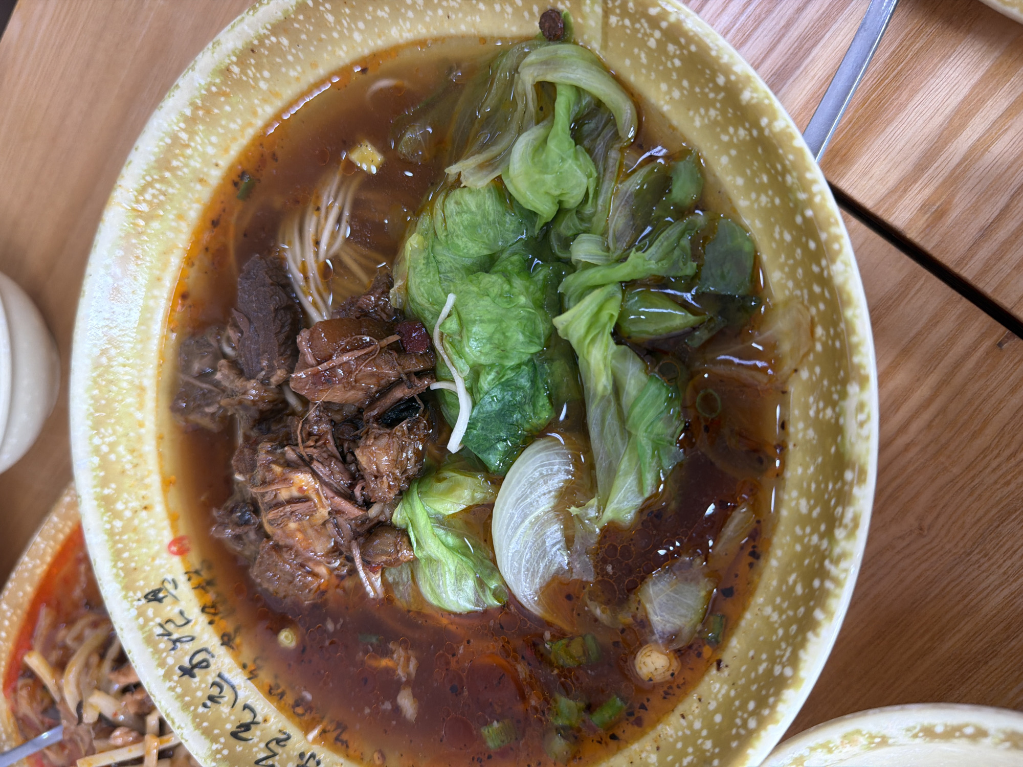 A close-up, overhead shot shows a bowl of noodles with braised beef and vegetables, likely in a restaurant setting. The bowl is yellowish with a speckled pattern and rests on a wooden table. The noodle soup is rich and red, filled with shredded beef, white noodles, and large pieces of lettuce and onion. Some black speckles, possibly chili flakes, are visible throughout the broth. Chinese characters are written along the rim of the bowl. The lighting is bright, suggesting an indoor environment. No specific location in Davao City, Philippines can be identified.