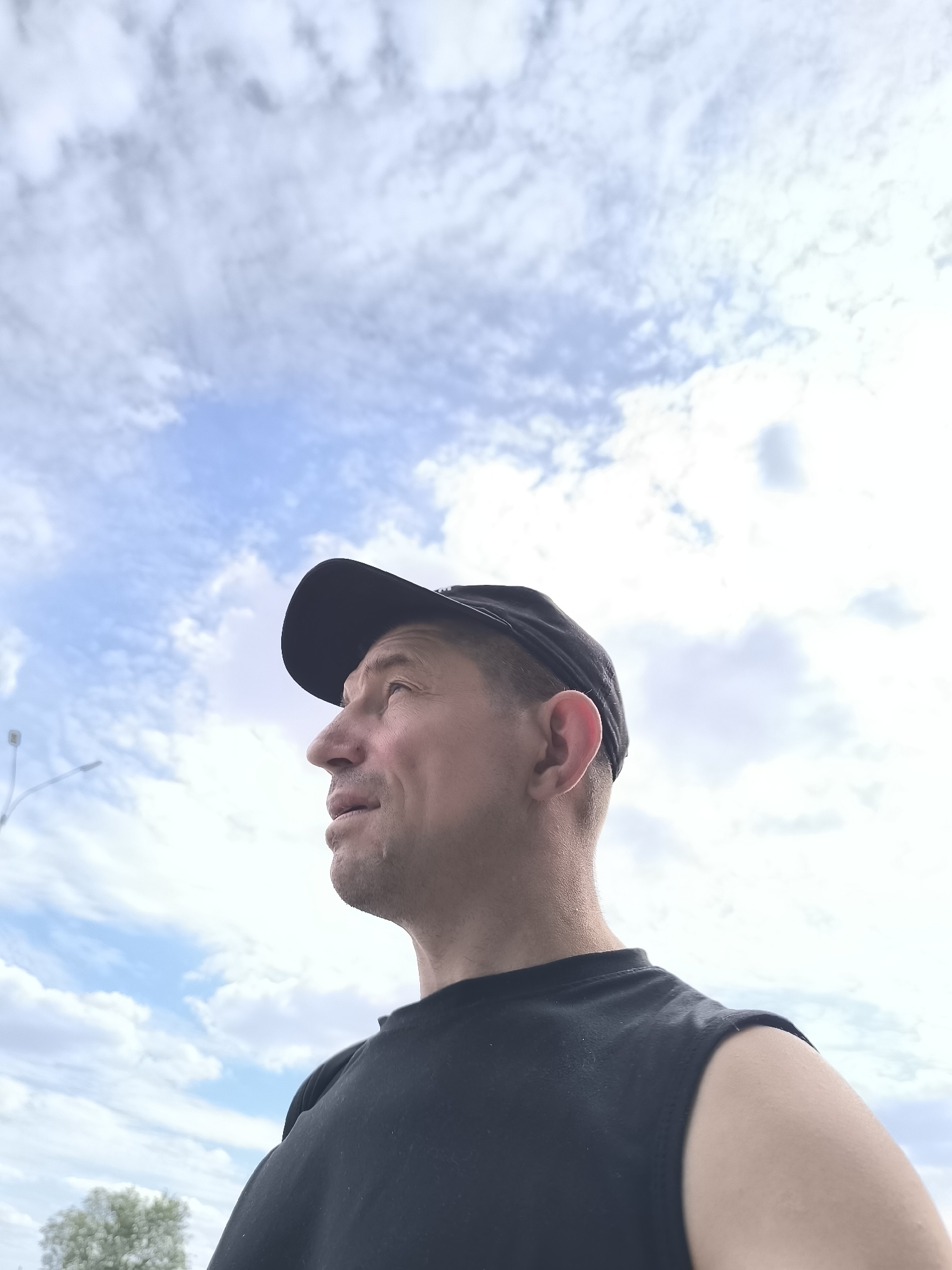 A low-angle, outdoor, waist-up shot features a Caucasian man from the chest up, facing left and looking upwards. He is wearing a black t-shirt and a black baseball cap. The background is a bright, overcast sky with scattered clouds. A faint street lamp is visible in the upper left portion of the image. The man's face is weathered, with visible pores and a slight sheen on his forehead. His lips are parted slightly, and his eyes are looking towards the sky. The man's skin tone is fair. The overall lighting suggests daytime. The image does not contain any readily identifiable landmarks or features that would definitively place the location within the city of Karaganda, Kazakhstan.