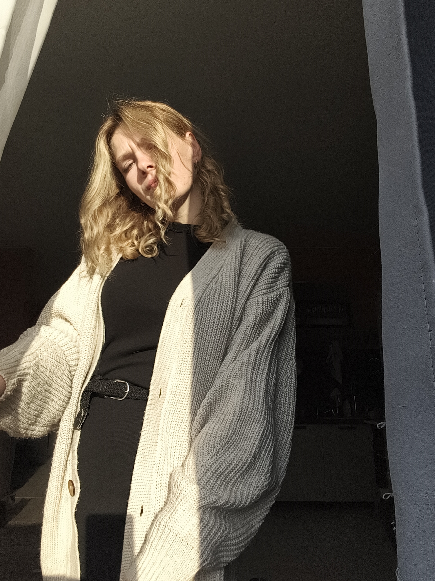 A young, light-skinned woman with wavy blonde hair stands in a shadowed doorway. She is wearing a black dress cinched with a black belt and a cream-colored knit cardigan. Bright sunlight illuminates part of her face and the cardigan. The background is dark, but a glimpse of a kitchen area with cabinets and possibly a figure can be seen. The location is unknown; there is no information in the image to suggest the location is Русский Вожой, Russia.