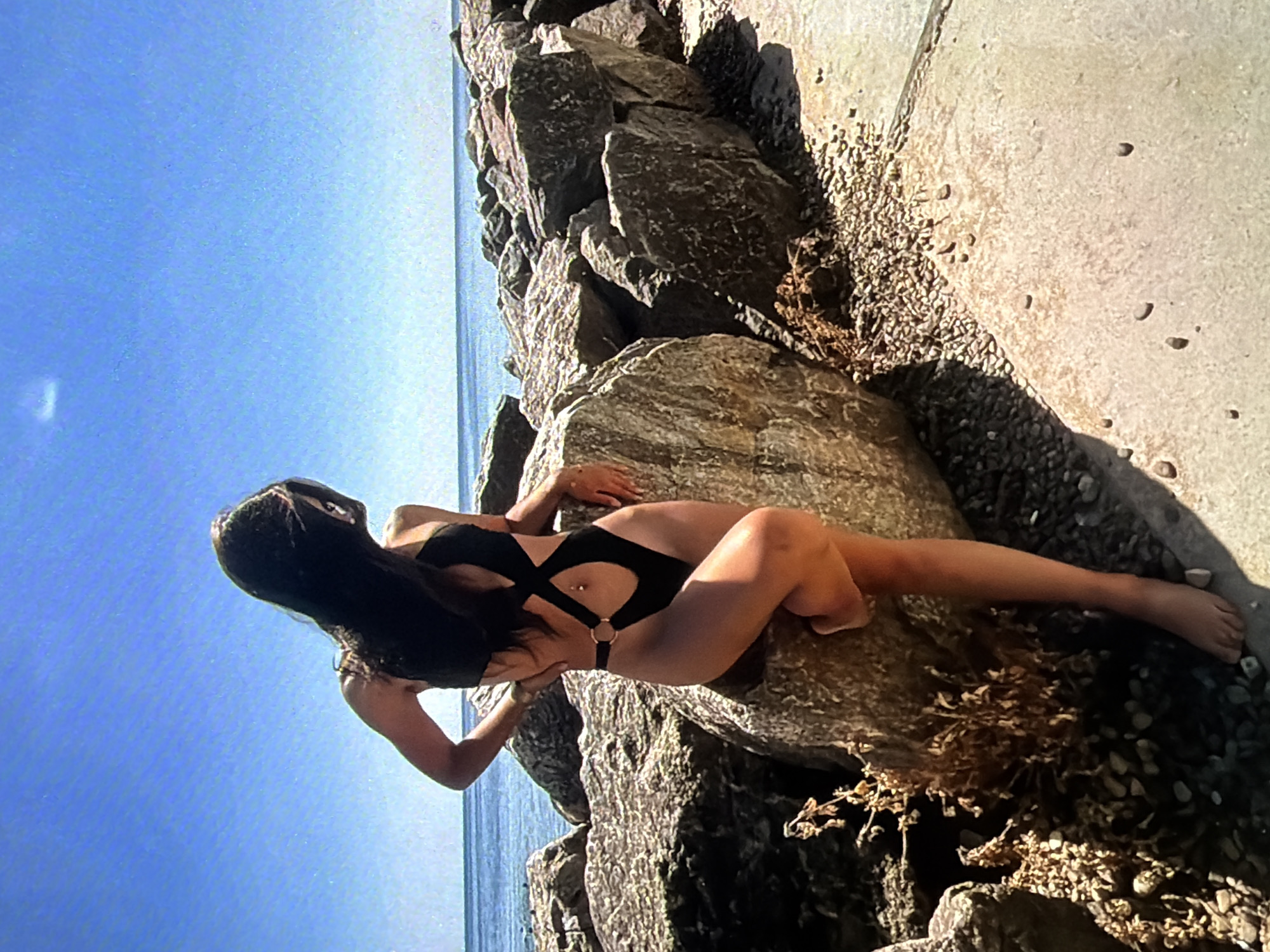 A woman with long dark hair, wearing a black one-piece swimsuit with cut-outs and a gold ring detail, is leaning against a large rock on a rocky shoreline. She is positioned to the left of the frame, with her body angled away from the camera, looking out towards the water. The sea is visible in the background, stretching to the horizon under a clear blue sky. The foreground consists of rocks and a paved walkway. The image is a full-body shot of the woman and her immediate surroundings. The location is not in Brunswick, Germany.