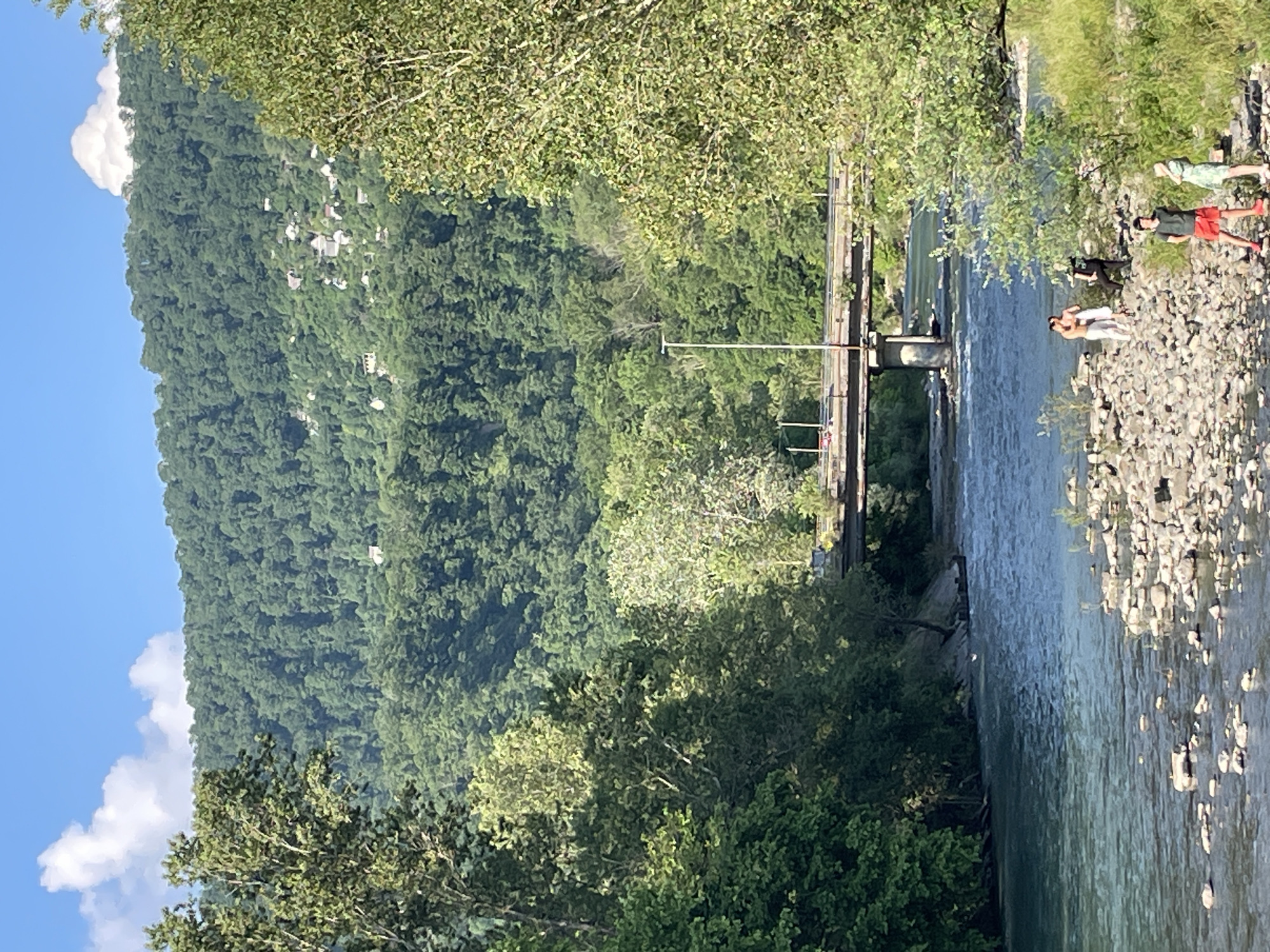 An eye-level, medium shot captures a serene river flowing through a lush green valley, with a bridge spanning the water in the mid-ground. The river, with its clear, turquoise water, is dotted with smooth, grey stones along its banks. The left bank is densely wooded with tall trees, while the right bank features a rocky shoreline where four individuals are present. Two individuals, a male and a female, stand close together, appearing to be in conversation. Another male is standing slightly behind them, looking towards the river. A fourth individual, a male in red shorts and a dark green t-shirt, stands in the foreground on the rocky bank, facing the camera. A woman in a light green dress stands to his right, looking away from the camera. In the background, a heavily forested mountain slopes upwards, with small houses and buildings scattered across its surface. The sky above is a clear blue, with a few fluffy white clouds. The bridge, constructed of concrete and steel, is supported by multiple pillars and has a road with a few vehicles visible on it. A single lamppost stands beside the road on the bridge. This scene is likely located in a mountainous region near Sochi, Russia, given the characteristic landscape.