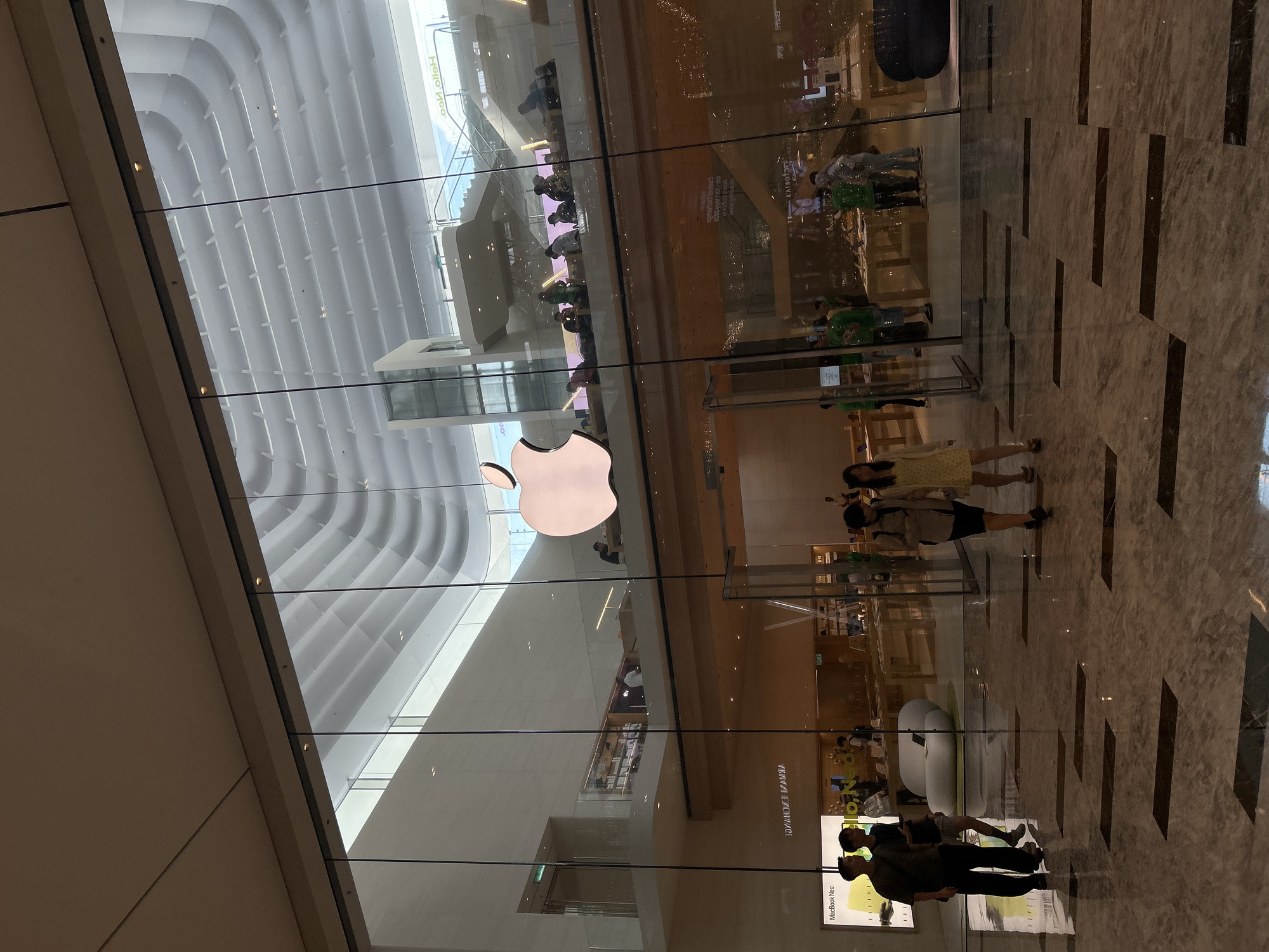 The image shows an Apple Store in Kuala Lumpur, Malaysia. The store has a large glass facade with the Apple logo prominently displayed. Inside, customers and Apple employees can be seen. The interior is well-lit, with displays of Apple products. The architecture is modern with distinct ceiling design. The floor is patterned with rectangular tiles. Outside the store, pedestrians are walking on the marble floor.