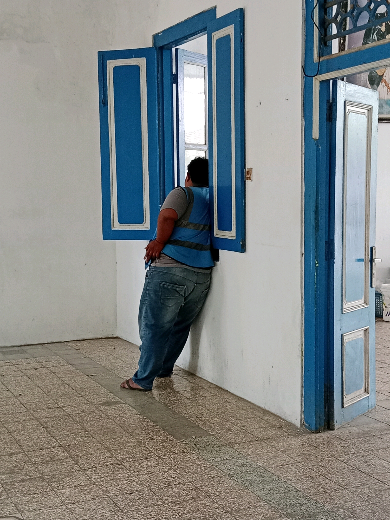 A person wearing a blue safety vest and denim jeans leans against a white wall, looking out an open blue-framed window. The window is divided by a white frame. To the right, a blue-framed door stands open, revealing more of the interior. The floor is tiled with a speckled pattern. The scene appears to be an interior space with white walls and a tiled floor, suggesting it could be a building or establishment within Jentekan, Indonesia.