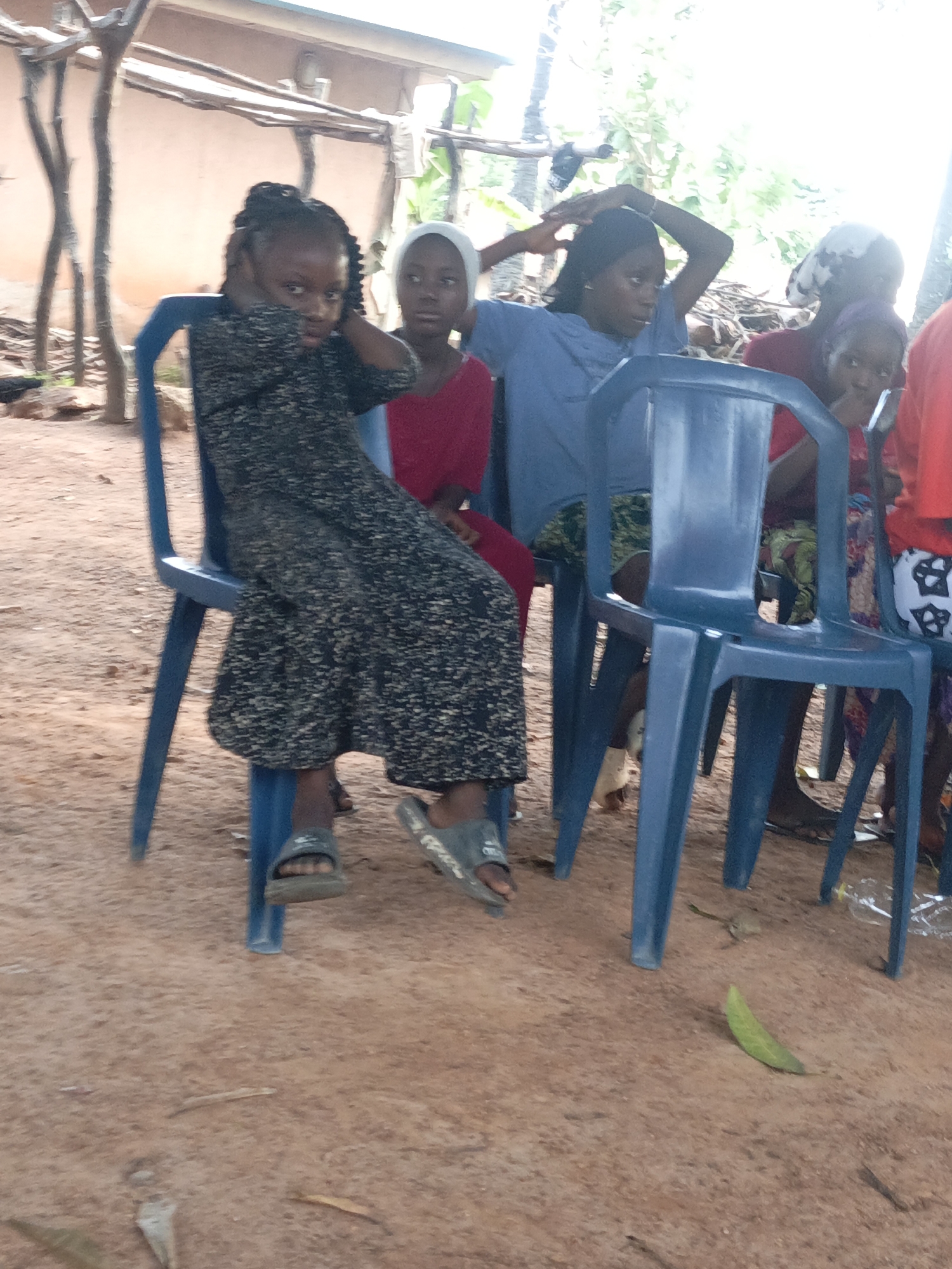 The image shows a group of young people seated on blue plastic chairs outdoors in Akwanga, Nigeria. The ground is reddish-brown earth with scattered leaves.
From left to right, the subjects are:
1. A young girl wearing a dark patterned dress and sandals, sitting in the foreground.
2. A girl in a red dress and a white head covering.
3. A girl in a blue shirt and patterned skirt, with her hands raised to her hair.
4. A girl in a red top and patterned leggings.
5. A girl in a red top, and partially visible on the right.
The background includes a building with a light-colored wall and a thatched roof structure. There is sparse vegetation visible in the background.