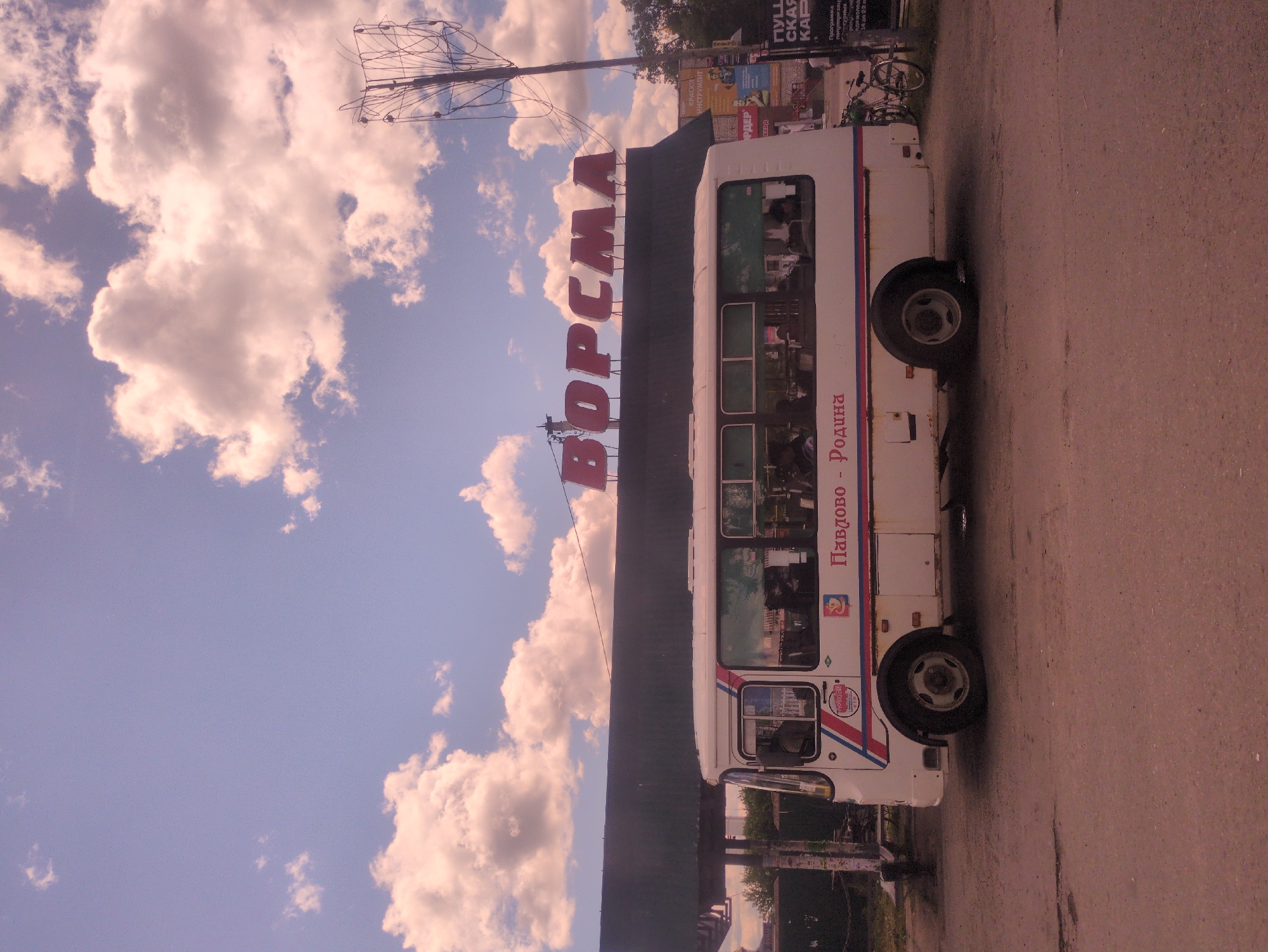 A white bus with red and blue trim is parked on a dirt road in front of a building with the word "ВОРСМА" (Vorsma) in large red letters across its facade. The bus has several windows, and people can be seen inside. The building appears to be a commercial establishment, possibly a bus station or a store. There is a telephone pole with wires on the right side of the image, and a bicycle is leaning against it. Several posters are visible on the wall of the building to the right of the bus. The sky is blue with scattered clouds, and the scene appears to be in an outdoor urban setting.