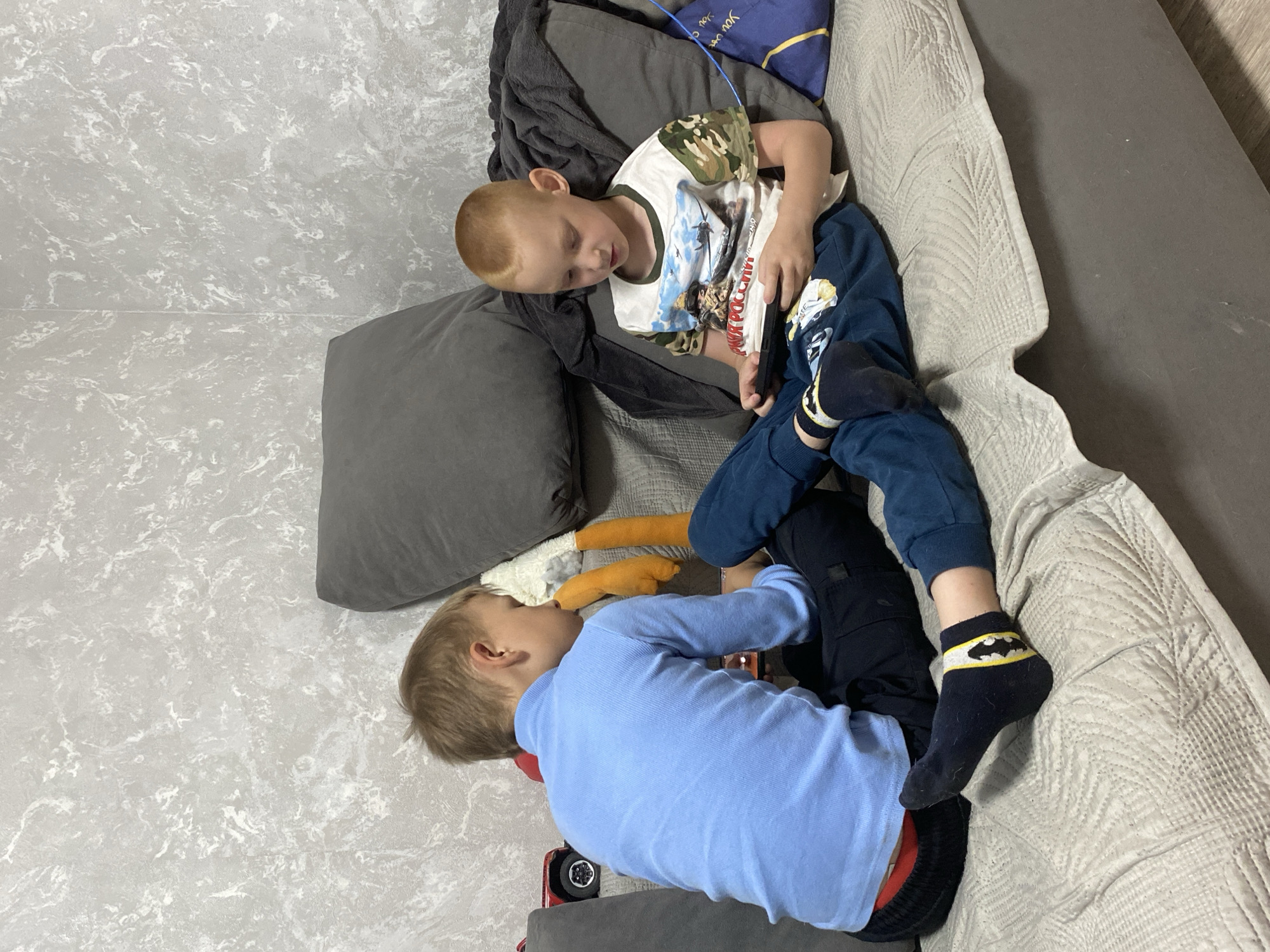 Two young children are lying down in a domestic setting. The child closest to the top of the frame is on a sofa, with a grey blanket, a navy blue pillow with gold lettering, and a grey pillow behind him. He is wearing a white, short-sleeved t-shirt with a camouflage pattern around the collar and sleeves, and the image of a fighter plane on the front. He is wearing dark blue pants and socks with a yellow design. The other child is lying on a grey tile floor. He is wearing a light blue long-sleeved shirt and black pants with a red and black striped edge. The socks are navy blue with a yellow bat design. A grey pillow is under his head and an orange and cream-colored stuffed animal is next to him. A small red toy is also visible. The furniture is against a light grey wall. The image was taken in Noviy, Russia.