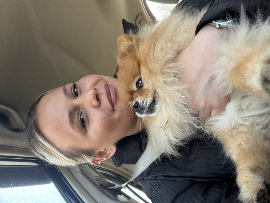 The scene is located inside a vehicle in the city of Makarovo, Russia. A female subject, with light blonde hair pulled back, occupies a passenger seat, facing the camera with a subtle smile. She wears a dark, textured jacket, secured by a light-colored seatbelt that crosses her torso. Her left ear displays multiple piercings: one helix, one tragus, and three lobe piercings. A silver-colored bracelet or watch is visible on her left wrist. She holds a small, fluffy Pomeranian dog, primarily reddish-blonde and cream in color, with both hands. The dog's head is slightly raised and turned towards the viewer's left, its dark eyes looking slightly upwards, and a small pink tongue is visible. The immediate environment is the interior of a vehicle, characterized by a light-colored headliner and a blurred window background indicating daytime outdoor conditions.
