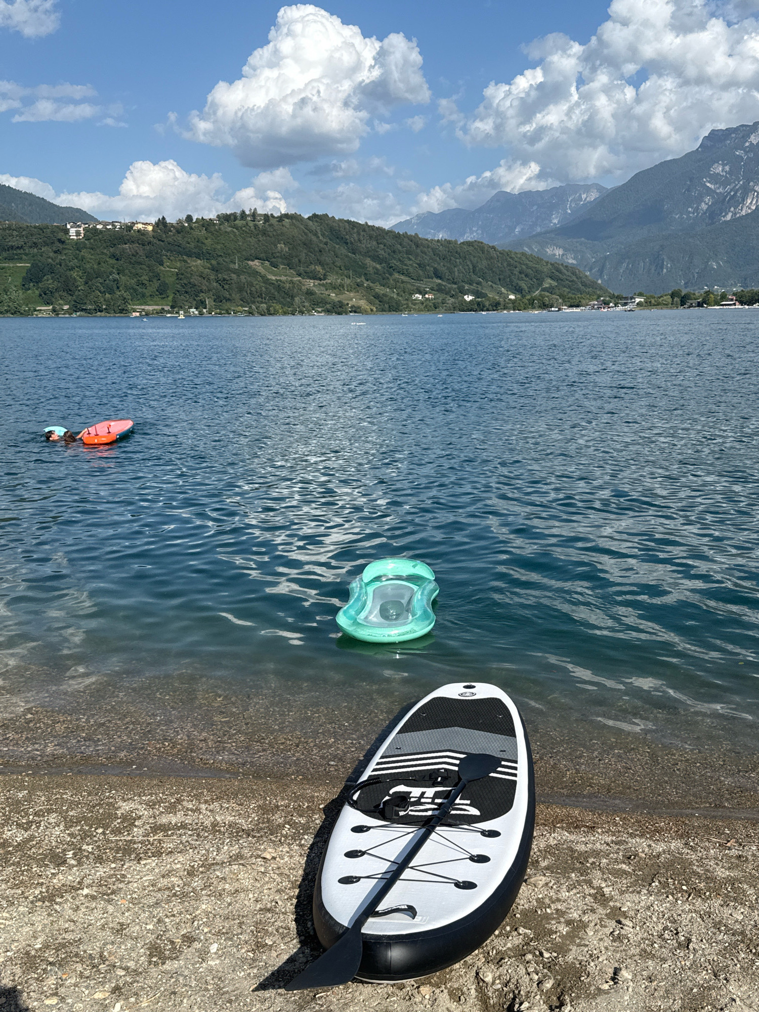 The image depicts a scenic view of Calceranica al Lago, Italy, on a bright, sunny day. In the foreground, a white and black paddleboard with a black paddle rests on a pebbly shore. The paddleboard is positioned at an angle, with the paddle lying across it. To the right of the paddleboard, the clear, blue water of the lake is visible, with small waves gently lapping the shore.
Further out in the lake, a green inflatable snorkeling mask floats on the water's surface. To the left of the mask, a person is partially submerged in the water, holding onto an orange inflatable mat. The person appears to be engaged in recreational activity.
In the mid-ground, the calm expanse of the lake stretches towards the opposite shore, where a lush, green hillside is dotted with houses and trees. The hillside rises towards a range of majestic, hazy mountains in the background, their peaks silhouetted against a bright blue sky filled with fluffy white clouds. The overall scene evokes a sense of tranquility and summer leisure.