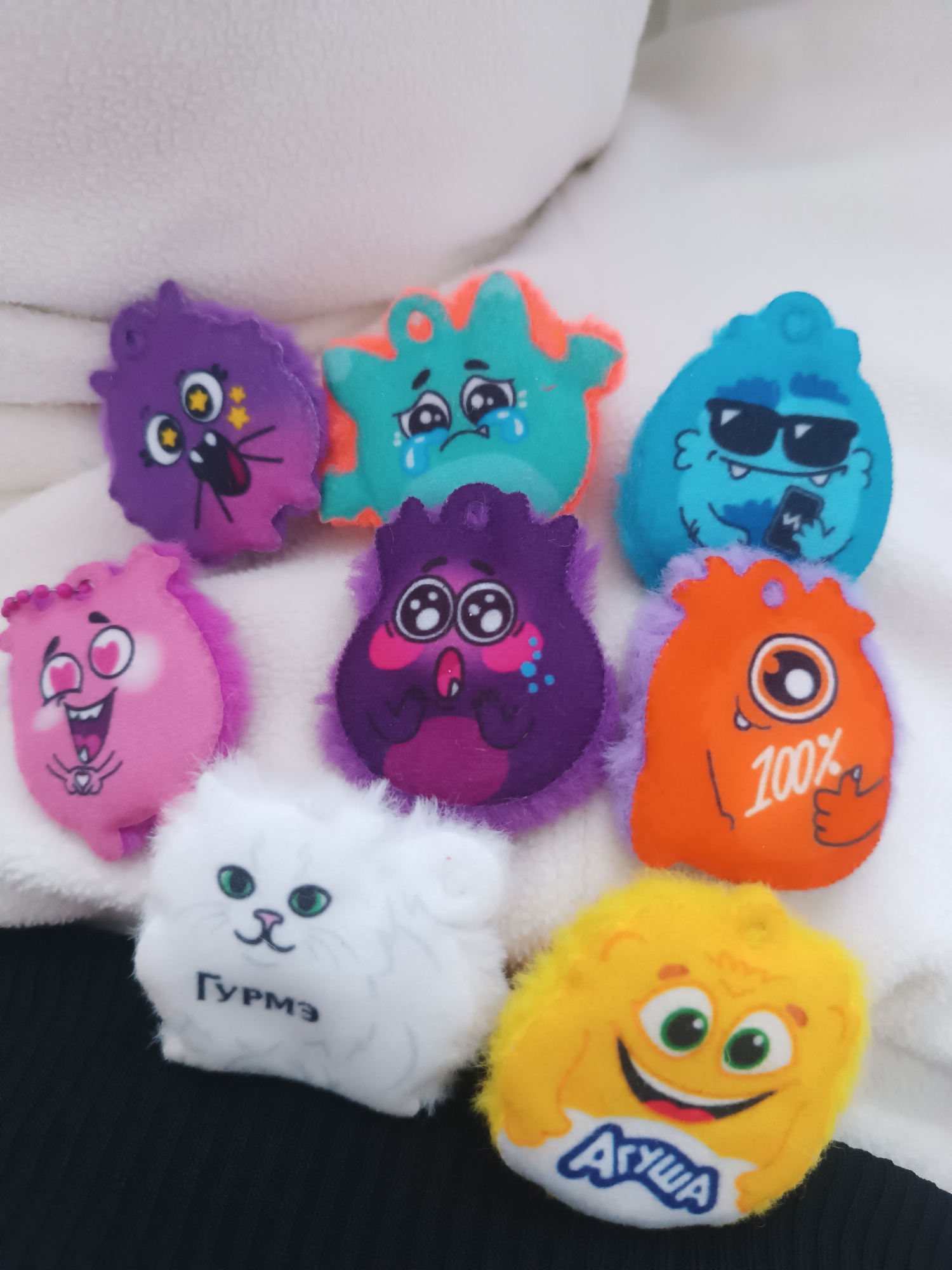 The image shows a collection of nine small plush toys arranged on a white, textured surface. At the top left is a purple monster toy with a surprised expression. To its right is a turquoise monster toy with orange accents and tearful eyes. Next to it is a blue monster toy wearing sunglasses and holding a smartphone. In the second row, starting from the left, is a pink monster toy with heart-shaped eyes and an open mouth. Adjacent to it is another purple monster toy with wide eyes and flushed cheeks. To its right is an orange monster toy with one eye and the inscription "100%". In the bottom row, on the left, is a white cat toy with green eyes and the Cyrillic inscription "ГУРМЭ". Next to it is a yellow monster toy with a smiling face and the Cyrillic inscription "АГУША". The toys are placed on a white, possibly furry or fleecy surface, and a black garment is visible in the bottom portion of the image. The location is unknown.
