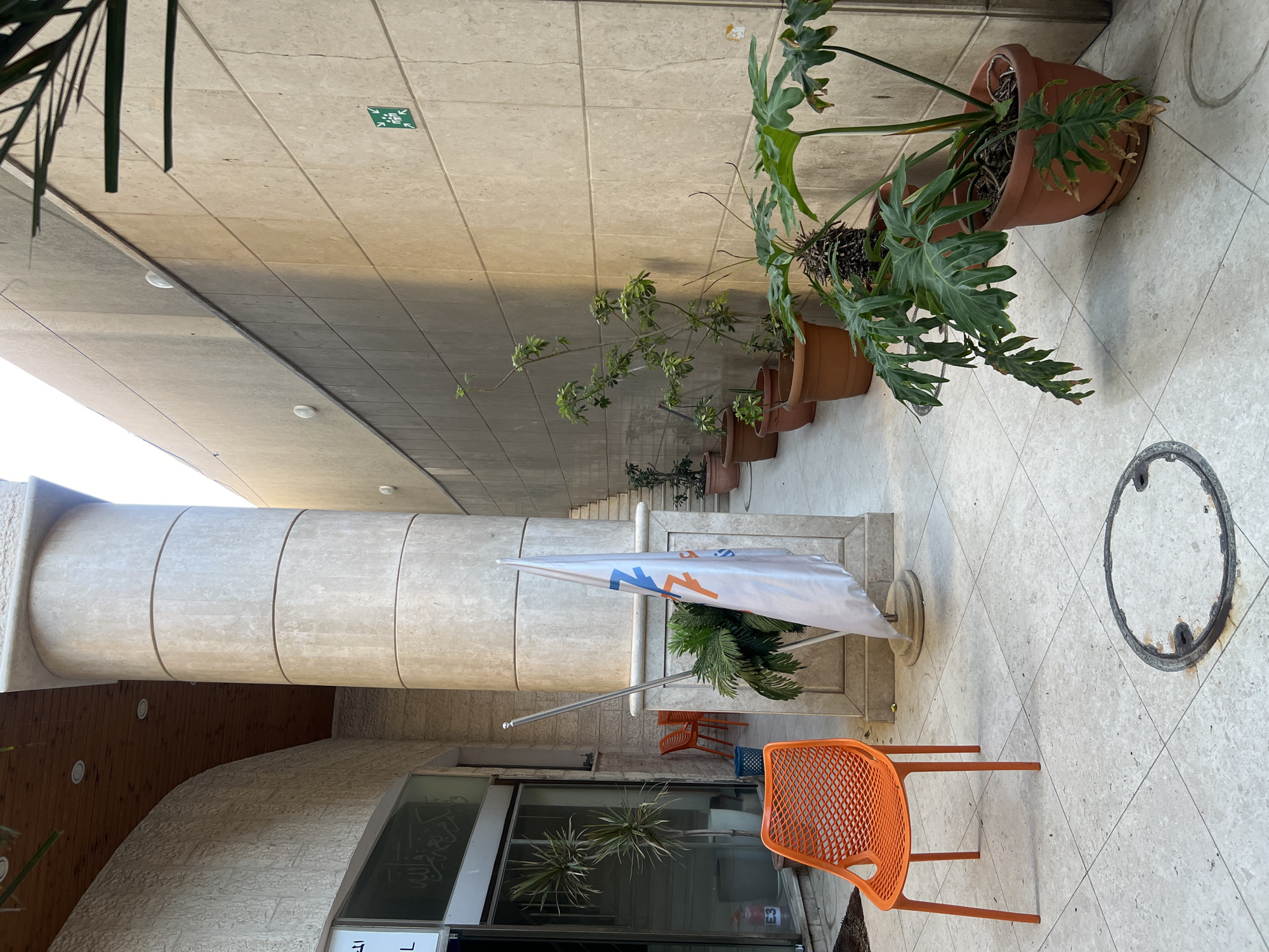 An outdoor paved walkway is depicted, with a large, light-colored cylindrical pillar on the left, separating the walkway from a building. The building has a curved facade on the left, with a wooden ceiling and recessed lighting visible. Further to the left, a glass-fronted entrance is visible, with Arabic script on the glass. An orange plastic chair with a mesh design is positioned on the walkway to the left of the pillar. Behind this chair, two more orange chairs and a blue bin are visible.
A white flag with orange and blue markings is planted on the walkway in front of the pillar. A large potted plant with broad, serrated leaves is to the right of the pillar. A row of potted plants in terracotta pots is lined up against a light-colored tiled wall on the right side of the walkway. The wall has horizontal grout lines. A green square sign with a white pictogram of people is affixed to the wall. In the foreground, a circular metal manhole cover is set into the paving stones. The scene is bathed in sunlight, casting shadows on the ground and wall. The overall impression is one of an accessible outdoor area, possibly a courtyard or entrance to a public building.