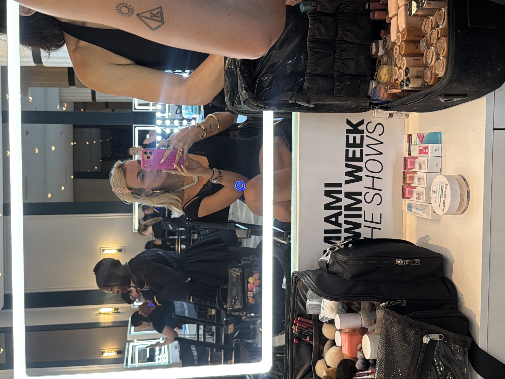 A mirror selfie captures a blond woman wearing a black dress and several bracelets and rings. She is holding a pink phone in her left hand. The mirror reflects the woman, and behind her are several makeup artists, who are also reflected. The environment appears to be backstage at an event, as indicated by the "Miami Swim Week" signage visible. Also visible on the table are several makeup products, cases, and brushes.