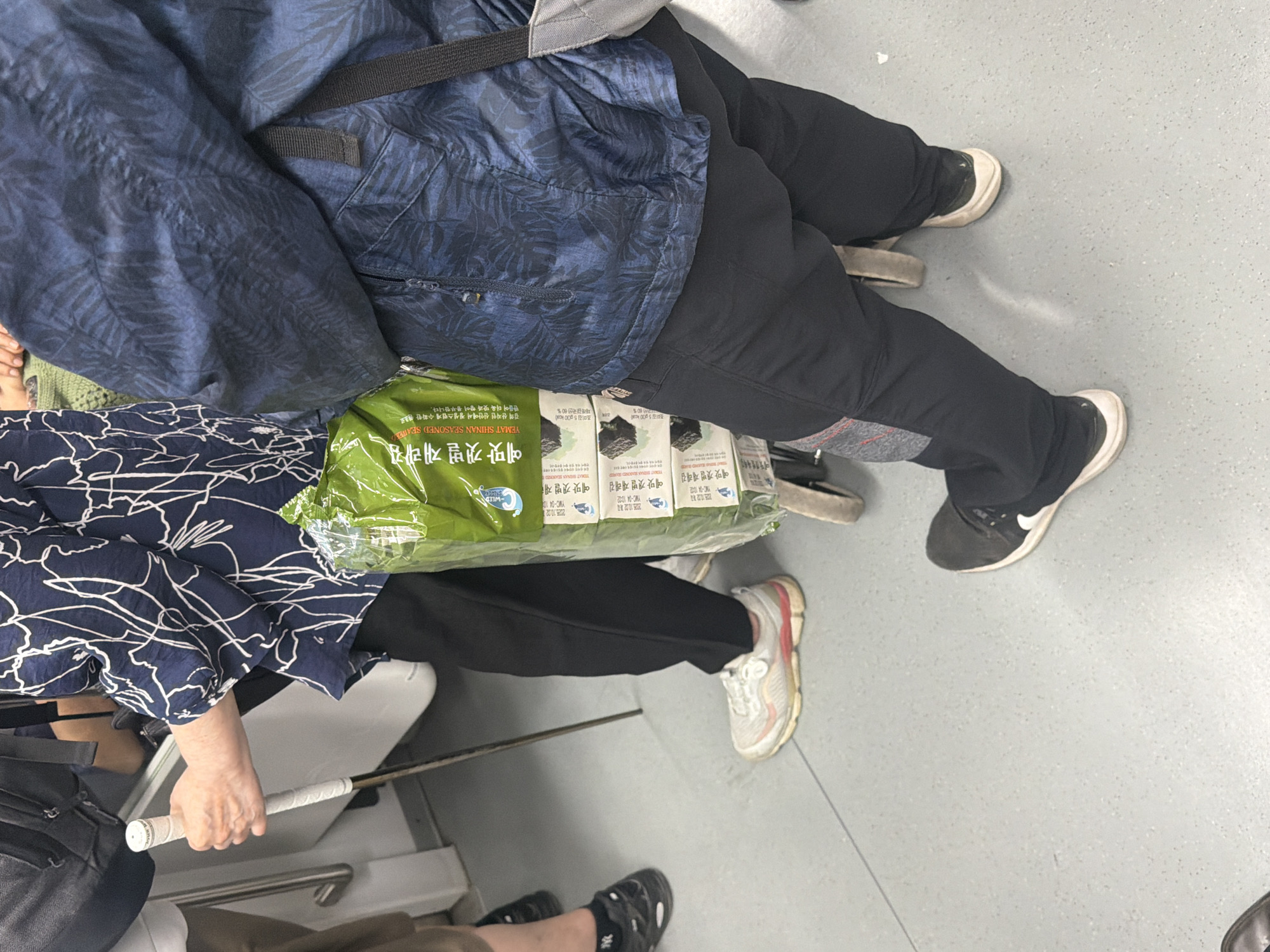 A high-angle shot shows two people sitting on a train, possibly in Seoul, South Korea. One person is wearing a blue leaf-patterned jacket and black pants, with a backpack slung over their shoulder. Their legs are stretched out, and they are wearing black and white sneakers. The other person is wearing a navy blue and white patterned top and black pants. They are holding a white golf club. A large package of "Yemat Shinan Seasoned Seaweed" rests between them. The train floor is gray.