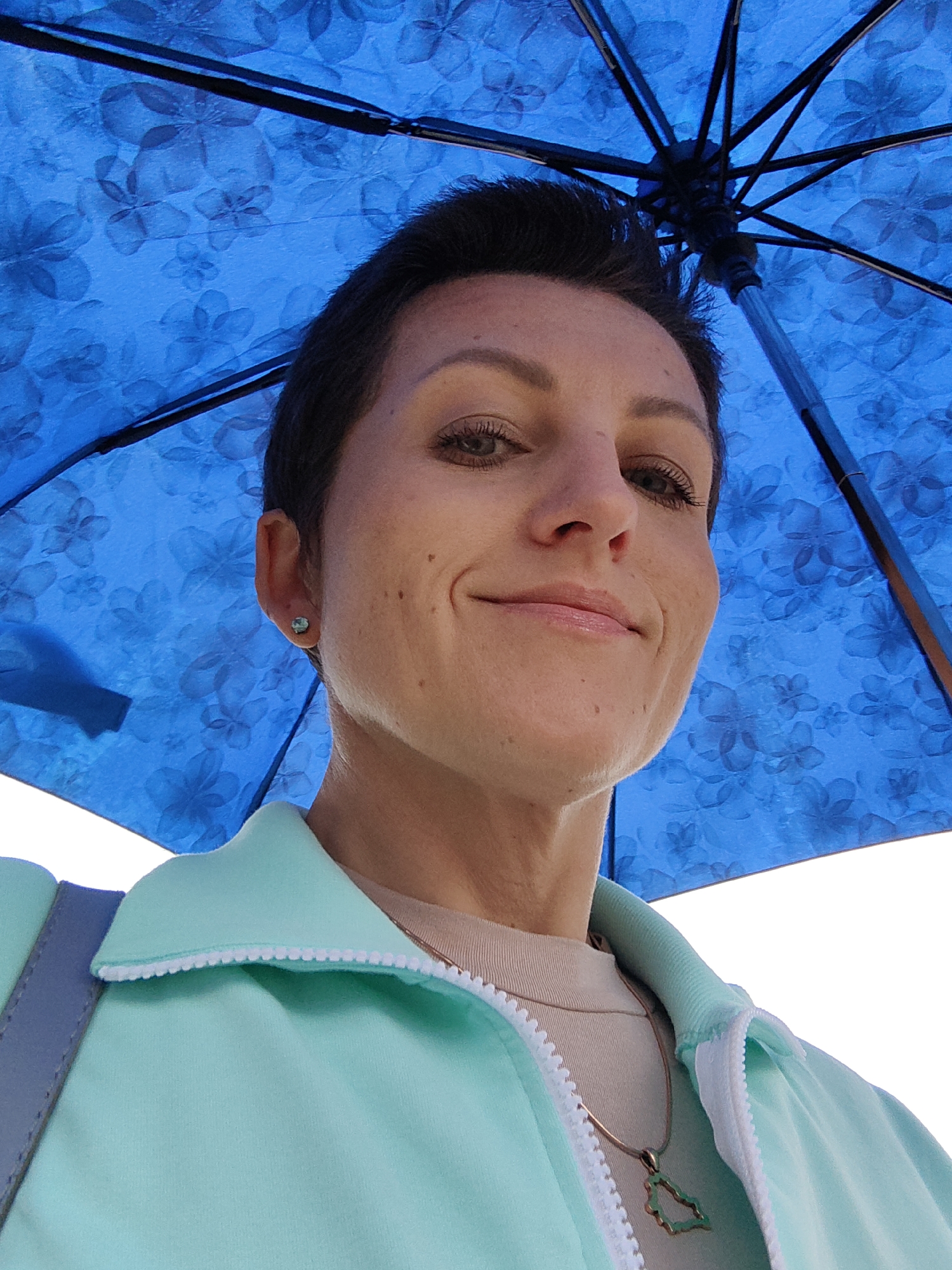 A low-angle, close-up, outdoor, waist-up shot captures a fair-skinned woman with short, dark hair and light brown eyes looking directly at the camera. She is wearing a light mint green zip-up jacket over a beige top, with a thin, gold chain and a pendant necklace. A small, aquamarine stud earring is visible in her left ear. The woman is positioned directly beneath a large, open blue umbrella with a floral pattern. The background is out of focus and appears to be a bright, white sky, suggesting an outdoor setting in Fanipal, Belarus. The image is well-lit, with the light source seemingly originating from above and slightly to the left, creating some shadows on the woman's face.