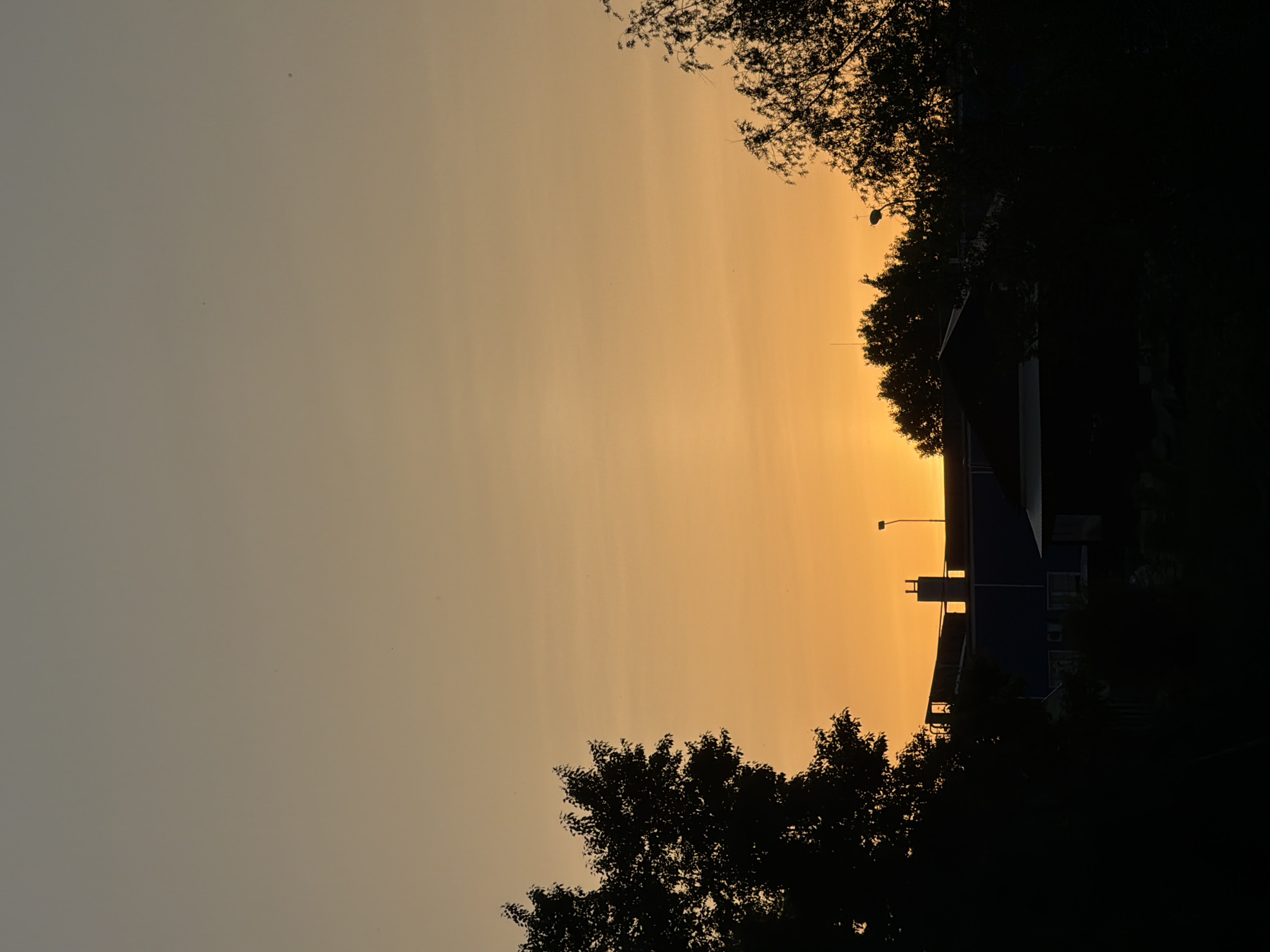 The image captures a sunset scene in Chaikovsky, Russia. The sky displays a gradient of colors, transitioning from a light grey on the left to a vibrant orange near the center. Silhouetted trees frame the top and bottom of the image. The dark outline of a building is visible amidst the trees, with a prominent chimney-like structure extending above its roof. A street lamp stands beside the building. The overall atmosphere is serene and still, emphasizing the natural beauty of the sunset against the urban landscape.