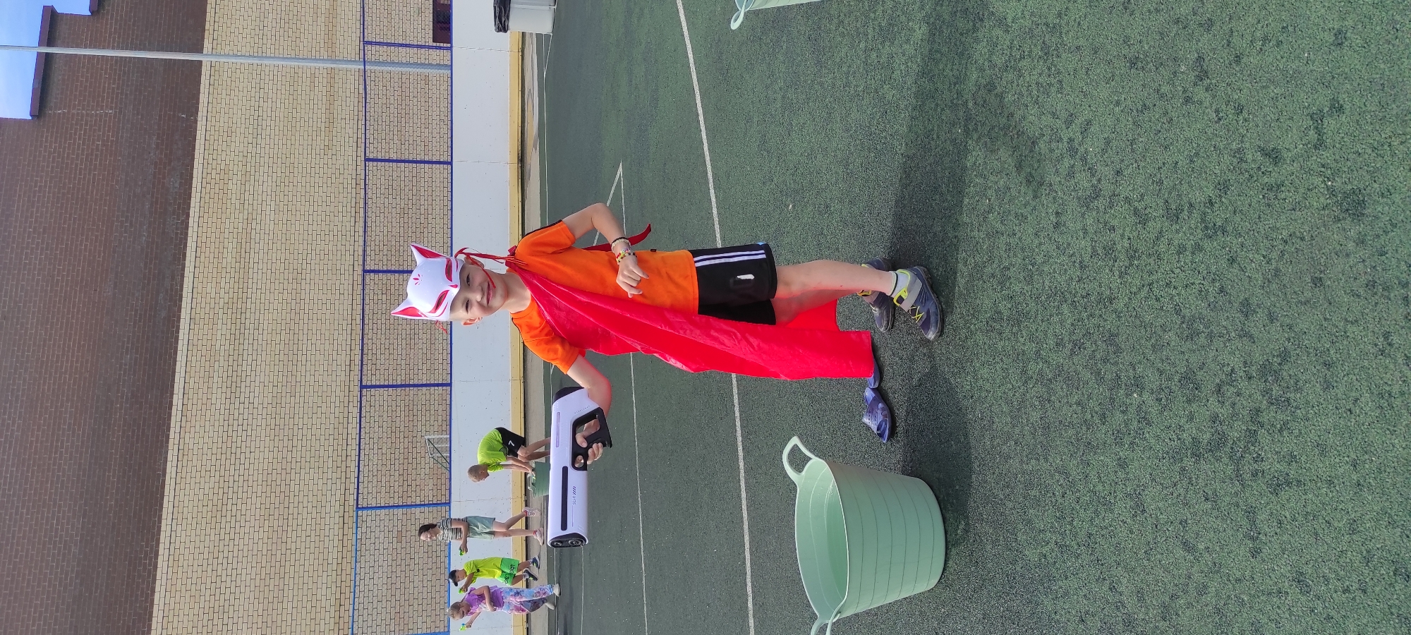 A young Caucasian boy, appearing to be around 7-9 years old, stands in the foreground on a dark green artificial turf field, possibly a sports pitch. He is wearing a white fox mask with red accents and strings, an orange t-shirt, black athletic shorts with white stripes, and grey and yellow sneakers. A bright red cape is draped over his shoulders. He is holding a white and grey water gun in his right hand, aimed slightly forward. To his left, a large, pale green plastic bucket filled with water sits on the turf. Further left and behind him, a group of children are visible. One girl in a pink top and patterned leggings is holding a water toy. Another boy in a bright green shirt is bending over a similar bucket. A girl in a striped t-shirt and shorts stands in the background, observing. The field is bordered by a blue fence and a brick wall. A grey pole, likely a light or flagpole, is visible on the right side of the image. The sky is a pale blue. This scene is likely at a public park or recreational area in Noviy, Russia, as indicated by the context of the request.