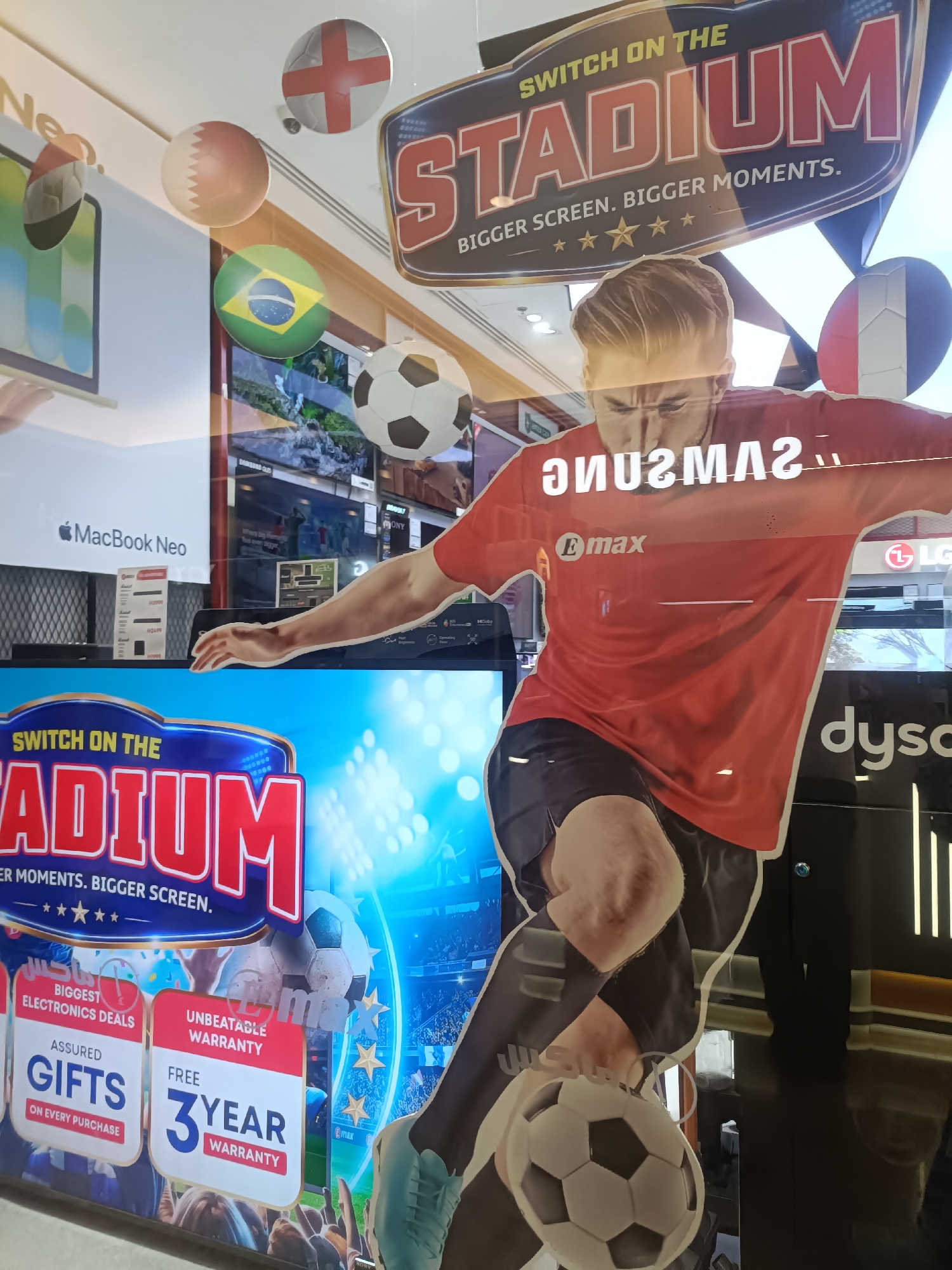 The image depicts a storefront display for electronics, likely in Dubai, United Arab Emirates, given the presence of flags of various nations commonly displayed at international events and the general aesthetic.
Dominating the foreground is a large, cut-out advertisement featuring a man in a red sports jersey, mid-stride, about to kick a soccer ball. The advertisement's text "SWITCH ON THE STADIUM" is prominently displayed in large, bold letters. Below this, smaller text reads "BIGGER SCREEN. BIGGER MOMENTS." and is accompanied by five yellow stars. The man's jersey reflects the text "SAMSUNG" in a mirrored fashion. There is also a logo that appears to be "Emax".
Hanging from the ceiling and interspersed within the display are several spherical objects resembling soccer balls, some adorned with national flags: England, Qatar, Brazil, and France.
To the left of the central figure, a MacBook Neo advertisement is visible. Further down and to the left, a large digital screen displays promotional graphics related to electronics deals, featuring text like "BIGGEST ELECTRONICS DEALS," "ASSURED GIFTS ON EVERY PURCHASE," and "UNBEATABLE WARRANTY," with a prominent "FREE 3 YEAR WARRANTY" offer.
The overall environment appears to be a retail space, with reflective surfaces suggesting a window or glass partition. The lighting is bright, and the background shows reflections of other retail displays and overhead lighting fixtures. The scene is busy, with multiple visual elements competing for attention, typical of a commercial setting aiming to attract customers.