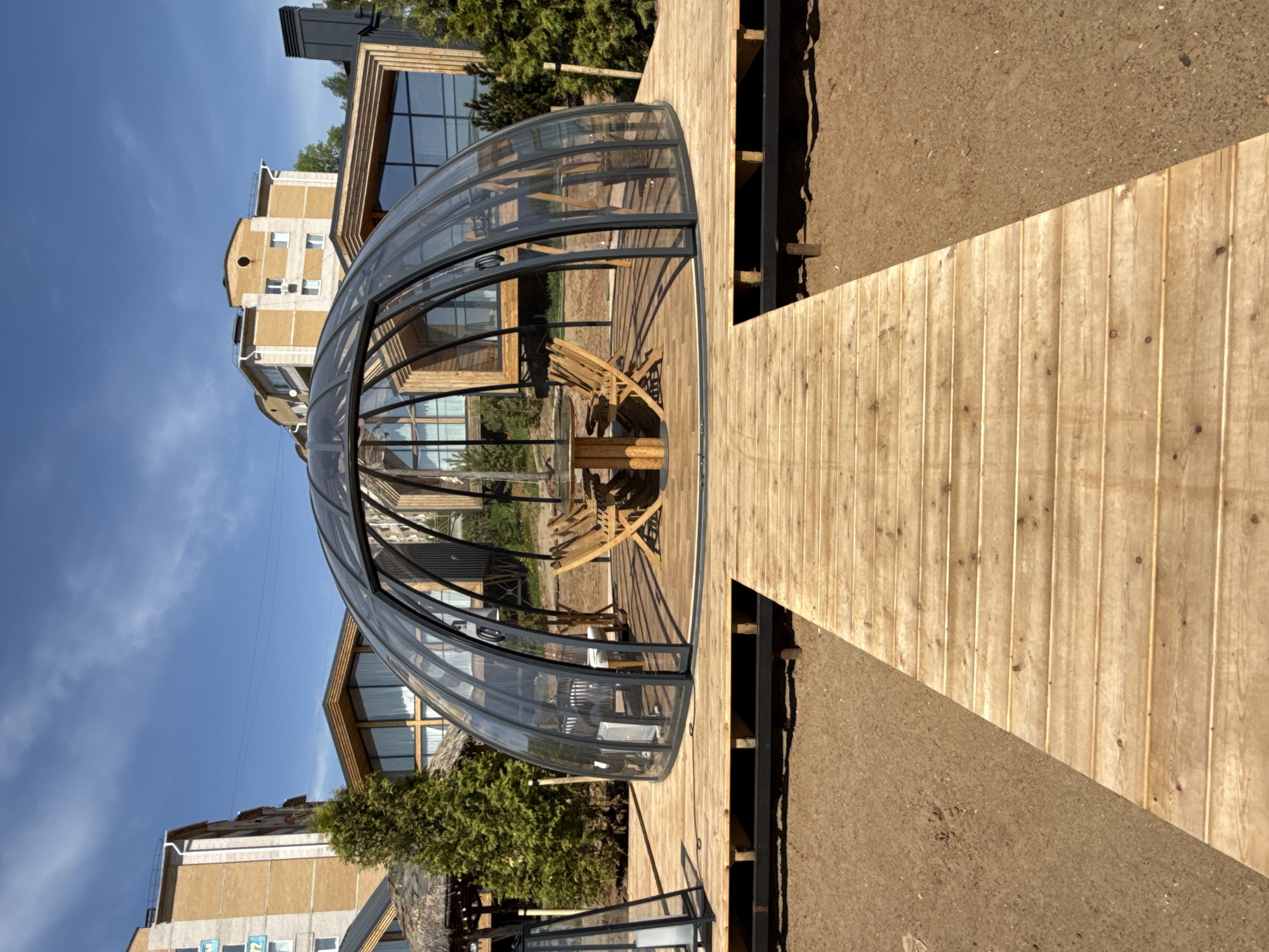 A curved glass enclosure stands on a wooden deck in what appears to be an outdoor setting, likely within the city of Votkinsk, Russia. The enclosure is rounded, almost dome-shaped, and it contains wooden furniture. The deck, made of light-colored planks, leads up to the enclosure and surrounds it. Beyond the deck, there is exposed ground or dirt. Buildings, some with modern architectural design elements, are visible in the background, along with trees. The sky is blue with some clouds.