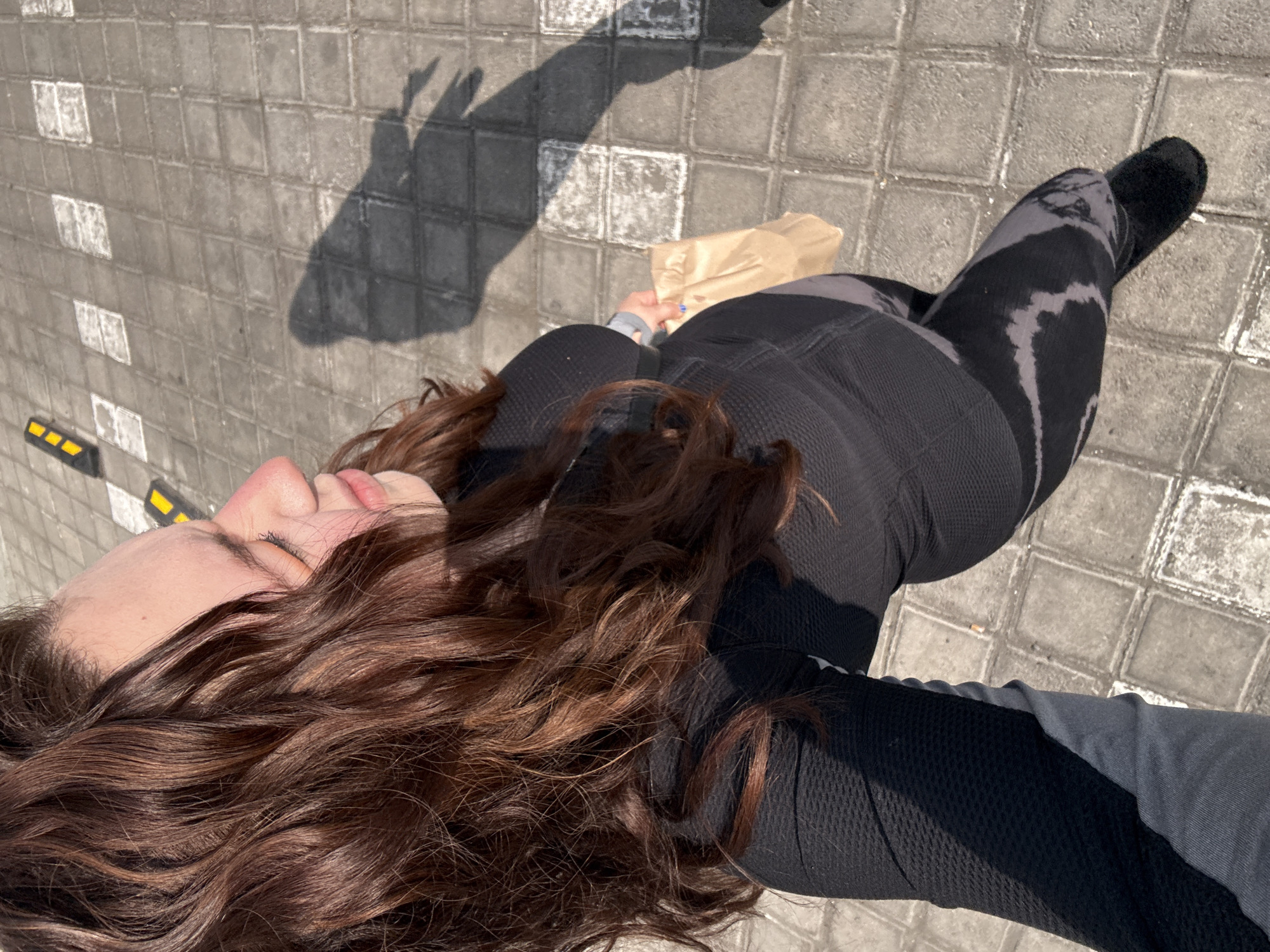 A person is standing on a tiled sidewalk. The person's face is visible, as well as long, curly, brown hair. The person is wearing black athletic clothing, consisting of leggings and a long-sleeved shirt. The right leg is also visible, and the person is wearing a black sock or slipper. In the background, the shadow of the person's hand is visible, forming a "peace" sign. The location of the image is Puebla, Mexico. A brown paper bag is being held in the person's left hand. The sidewalk tiles are grey and white.