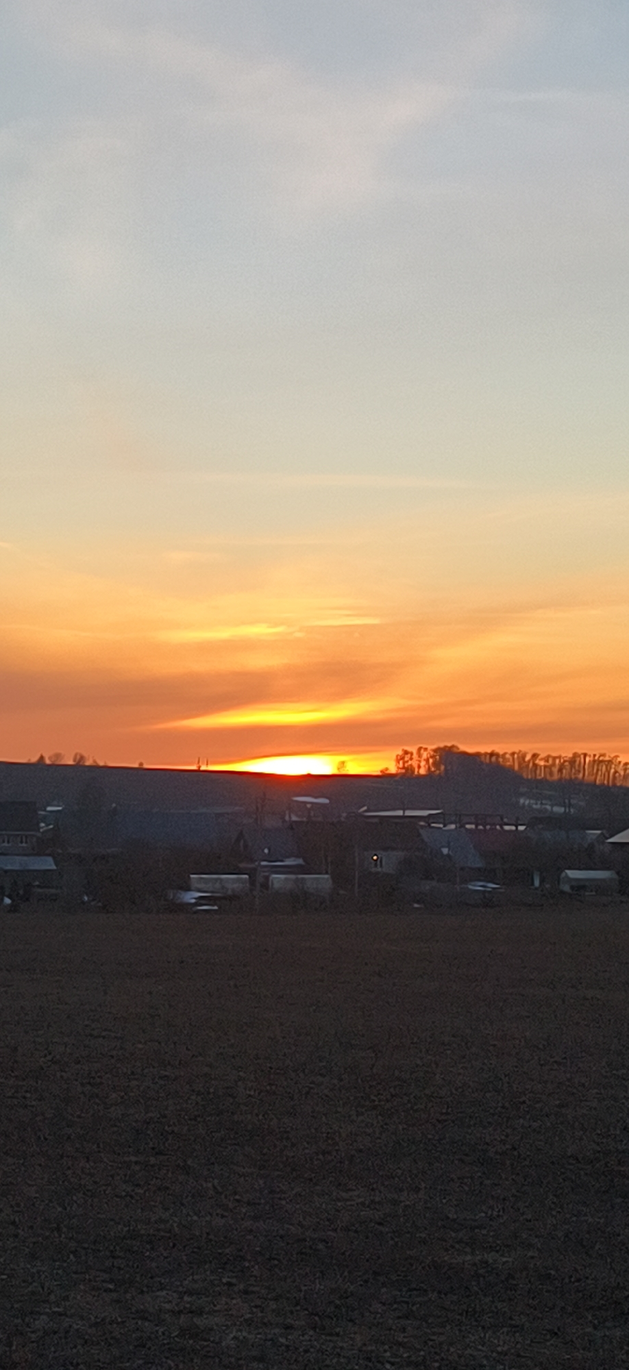 The image shows a sunset over a rural area, possibly near the city of Фоки, Russia. The sky transitions from light blue at the top to vibrant orange near the horizon where the sun is setting. A dark, ploughed field occupies the foreground, with a line of buildings and trees silhouetted against the bright sky in the middle ground. The buildings appear to be residential, possibly part of a small village or settlement. The overall impression is one of tranquility and rural landscape at dusk.