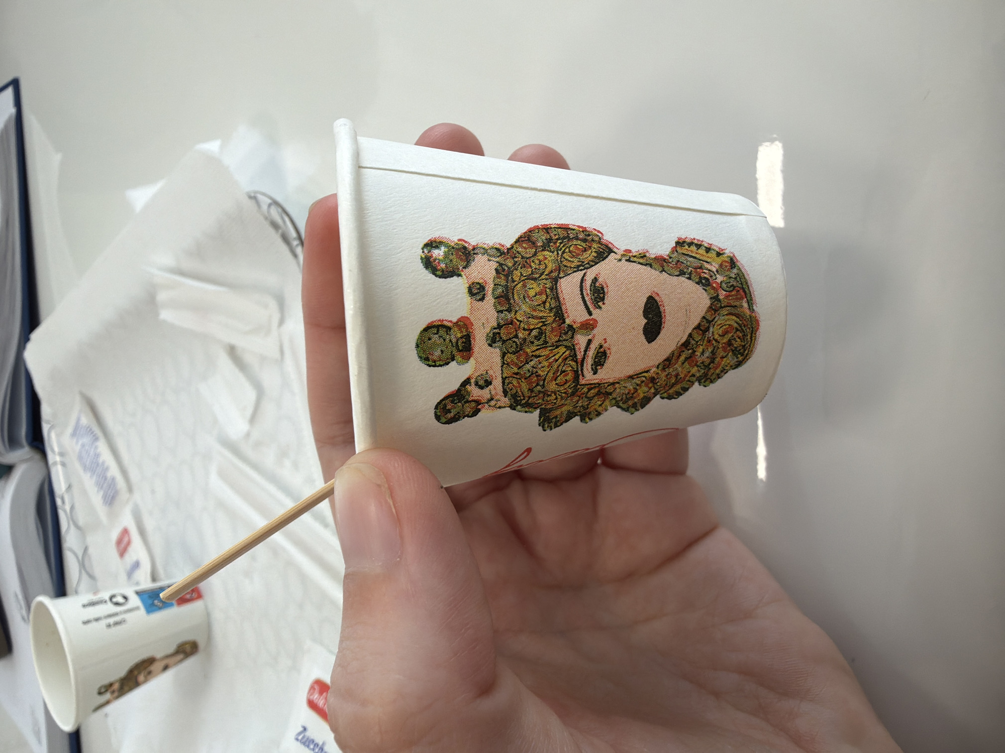 A person's hand holds a small white paper cup with a cartoon drawing of a person in ornate clothing. A wooden toothpick is inserted into the top rim of the cup. A second similar paper cup is on a white surface, which has packaging with the word "Zucchero". A white book is in the background. The location is Scordia, Italy.