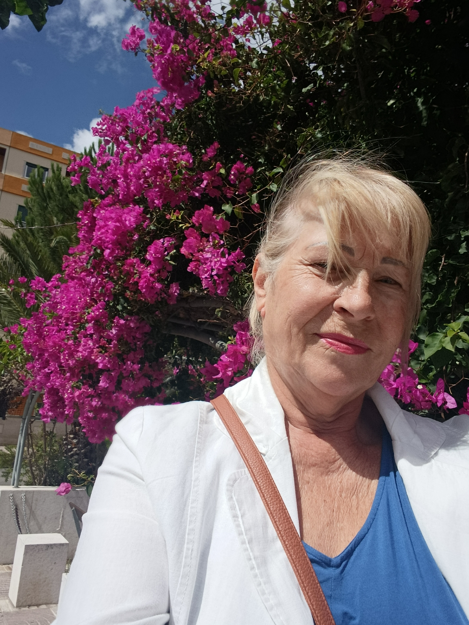 A woman with blonde hair, wearing a white jacket over a blue top, poses for a selfie in front of a vibrant pink bougainvillea bush. She is looking towards the camera with a slight smile. The background includes a building with orange trim and a blue sky with scattered clouds. The scene appears to be outdoors, possibly in a public space within the city of Setúbal, Portugal, given the presence of the characteristic bougainvillea and Mediterranean architectural style of the building.