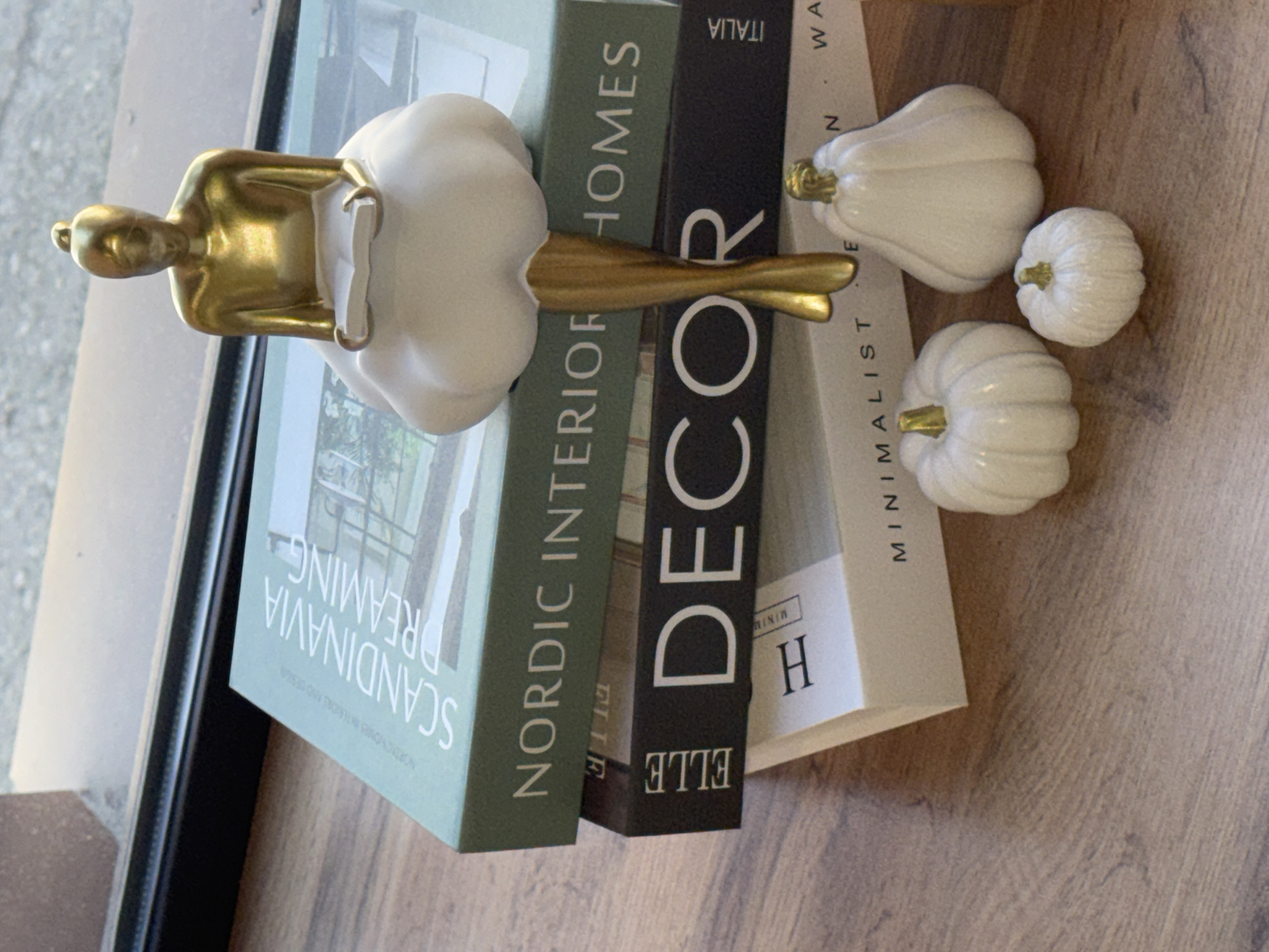 The image shows a stack of books and decorative objects on a wooden surface. The books are stacked horizontally, with the spines visible. The titles of the books are "SCANDINAVIA DREAMING," "NORDIC INTERIOR HOMES," "DECOR ELLE," and "MINIMALIST." On top of the books, a decorative figurine of a person reading a book. Three small, white pumpkin-shaped objects are next to the books. The scene is well-lit, and the colors are neutral and muted. The objects are likely arranged as part of a decorative display. The city of Chaikovsky, Russia is not visible.