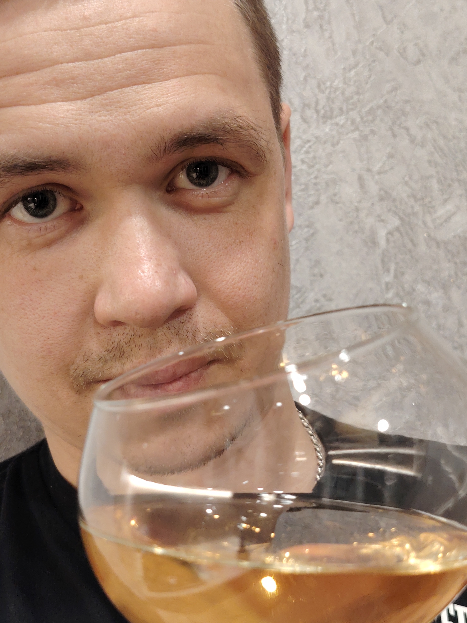 A close-up indoor shot shows a fair-skinned man holding a glass of amber liquid. The man's face fills the upper left of the frame, showing his eyes, nose, and part of his mouth and mustache. He has short brown hair and is wearing a black shirt. A glass containing a translucent amber liquid obscures the lower right of the frame. The background is a gray textured wall. There is no indication of the city or the fact that the city is called Chaikovsky.