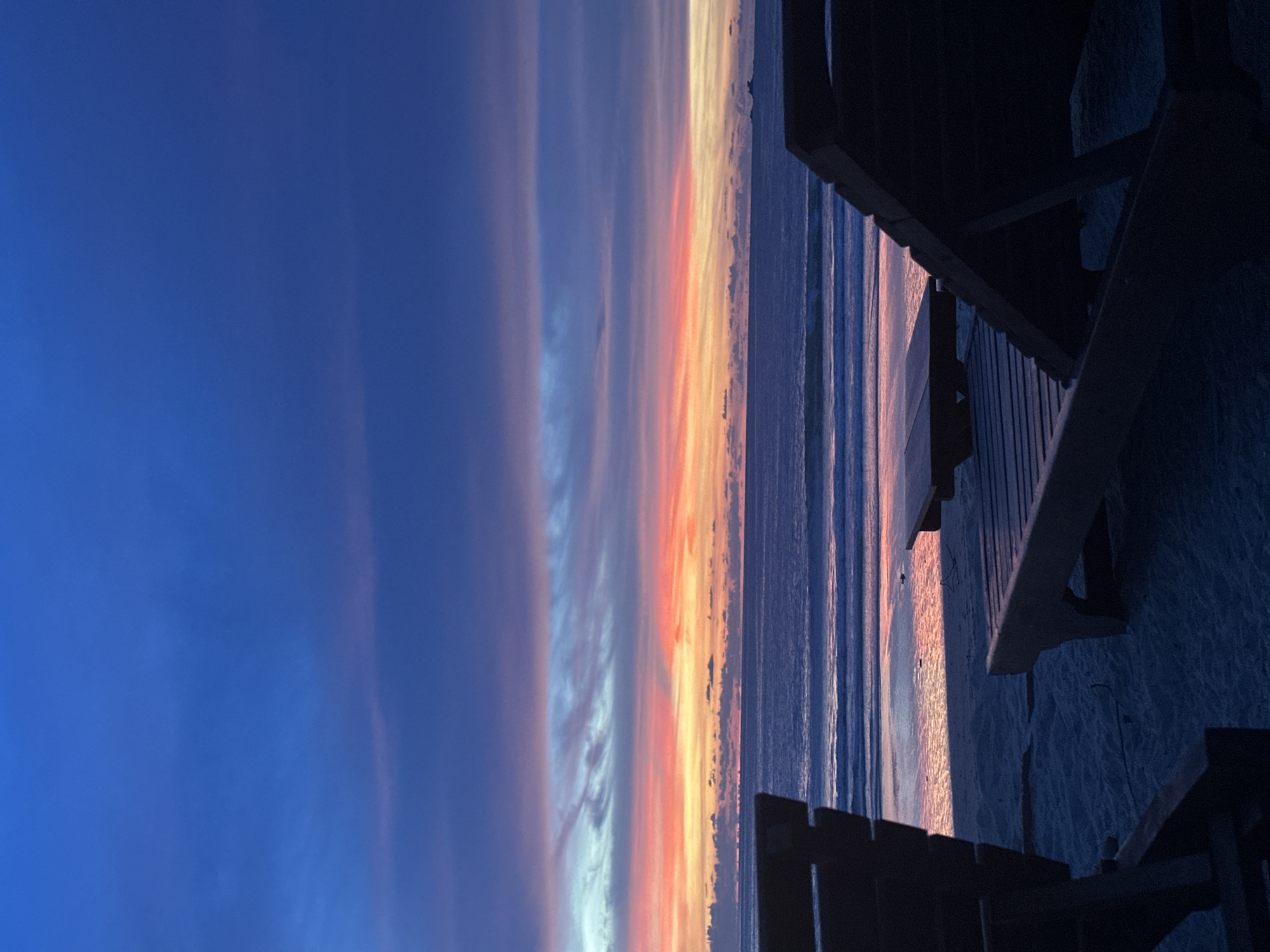 The image shows a beach scene at dusk. In the foreground, two dark-colored lounge chairs and a low table are silhouetted against the sand. The ocean stretches out in the midground, with gentle waves rolling towards the shore. The water reflects the colors of the sky. The sky itself is a gradient of deep blue at the top, transitioning to bands of pink, orange, and yellow clouds near the horizon. There is a faint outline of land on the far right horizon. The overall impression is one of tranquility and the end of a day.