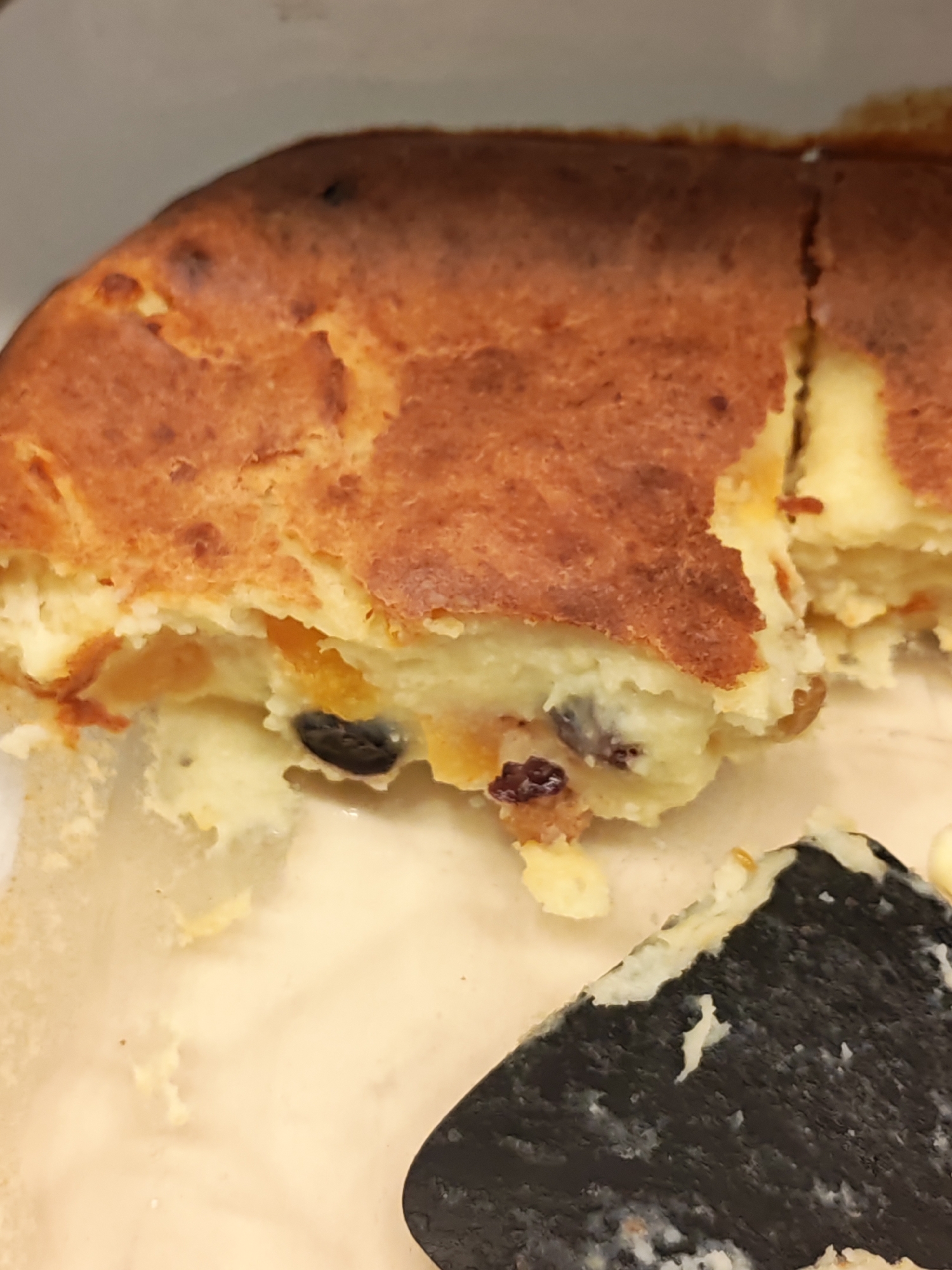 A close-up, overhead shot shows a golden-brown baked casserole with dried fruit inclusions. The casserole appears to be cut, revealing a creamy interior with visible raisins and apricot pieces. A black utensil is partially visible in the lower right corner of the frame. The scene is indoors, with a light-colored surface beneath the casserole and a pale gray background. There is no information in the image that relates to Bataysk, Russia.