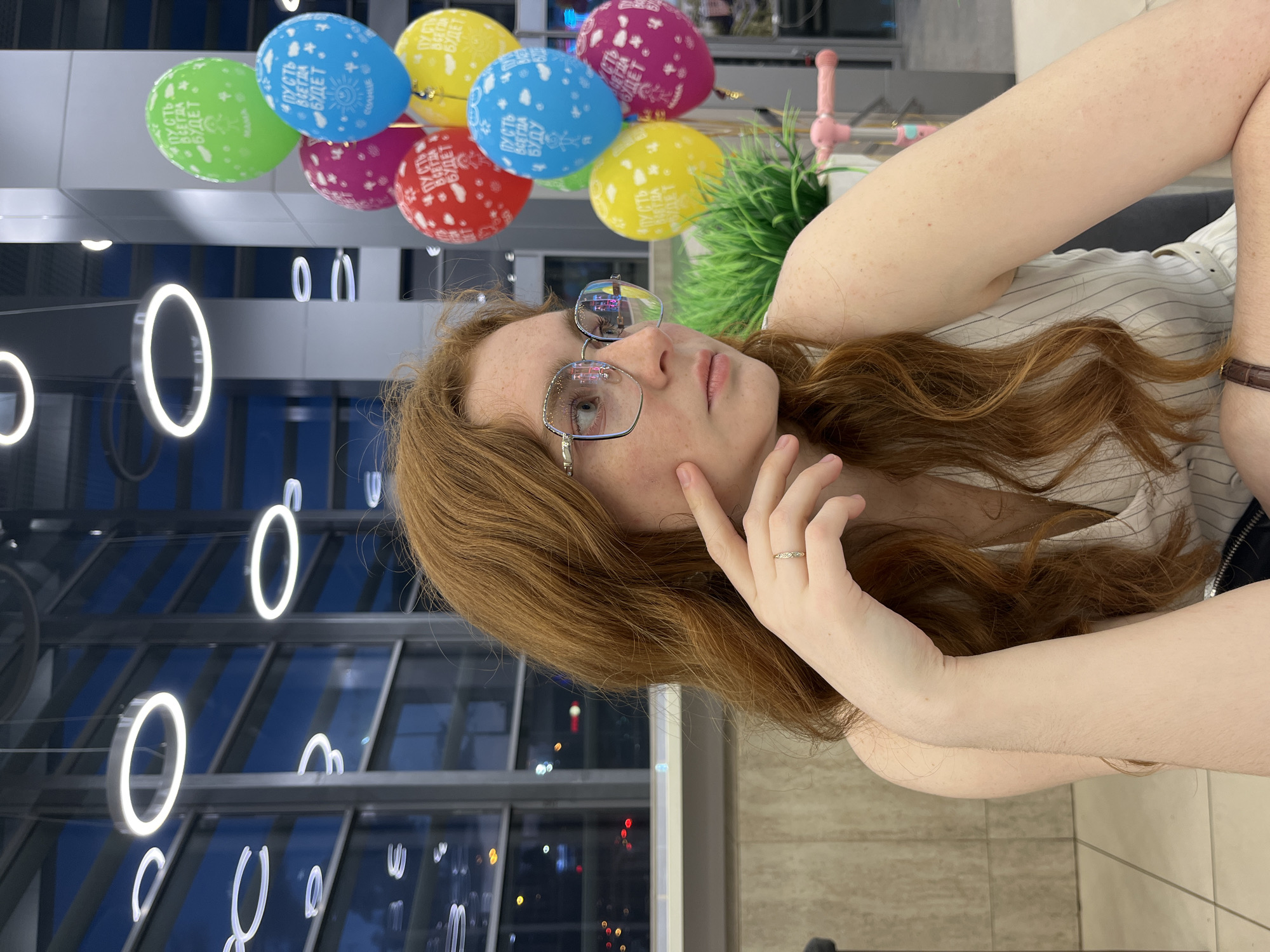 This portrait-style photograph features a young woman resting indoors, likely in Kazan, Russia. She has long, wavy reddish-brown hair, fair skin with freckles, and wears clear-rimmed glasses that reflect the ambient lights. Her expression is pensive as she looks slightly upwards and to her right, with one hand gently touching her face. She is wearing a light-colored, possibly striped, top, and a dark strap (perhaps from a watch) is visible on her right wrist.
Above and behind her float several colorful balloons in green, yellow, blue, pink, and red. White Cyrillic text is printed on them, conveying positive messages: a green balloon reads "ПУСТЬ ВСЕГДА БУДЕТ MAMA" (May there always be Mama), a blue one "ПУСТЬ ВСЕГДА БУДЕТ СОЛНЦЕ" (May there always be Sun), and other balloons display "ПУСТЬ ВСЕГДА БУДЕТ" (May there always be).
The background reveals a modern indoor environment, characteristic of a commercial building or public hall. Prominent features include large glass panels, through which dark outdoor scenes with city lights are reflected, suggesting it is evening or night. White, circular, and arc-shaped light fixtures are suspended from the ceiling. A decorative green plant and a light pink, potentially novelty, item are also visible. The overall scene combines personal reflection with a subtly festive atmosphere.