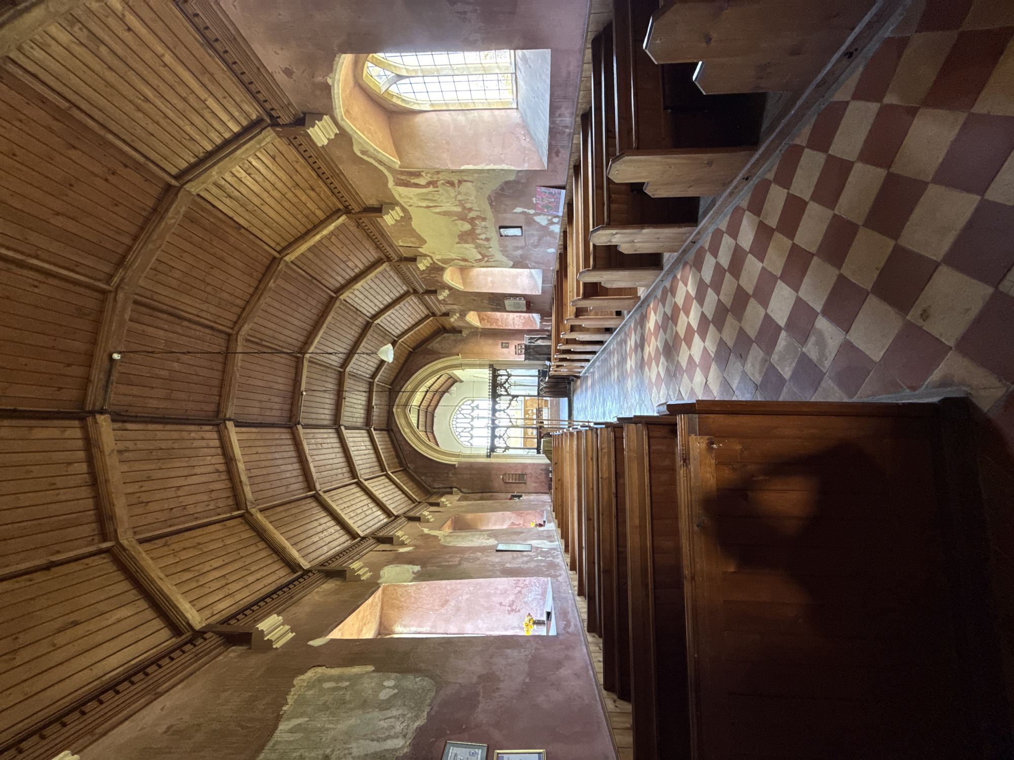 This media file captures the interior of a long, narrow hall, strongly suggesting a church or chapel in Great Yarmouth, United Kingdom. The perspective looks down the length of the hall, with natural light illuminating the scene.
On the left side of the image, an elaborate wooden vaulted ceiling curves upwards, constructed from numerous radiating panels and robust beams that form a fan-like pattern. Below this, a wall runs along the length of the hall, characterized by its distressed appearance with peeling reddish-brown paint revealing lighter, possibly cream-colored, layers underneath. Several rectangular window alcoves are set into this wall at a higher level, allowing bright daylight to stream in. Some of these alcoves contain small, indistinct objects.
On the right side of the hall, rows of dark wooden pews or benches are neatly arranged, indicating seating for congregants. Adjacent to the pews, the floor displays a distinct checkered pattern of alternating light and dark reddish-brown tiles. At the far end of the hall, a large, ornate arched window with intricate tracery serves as a prominent focal point.
The interior is brightly lit by natural light, casting some shadows, particularly on the wooden pews. No people or specific activities are depicted, giving the space a quiet, historical ambiance. There is a small framed picture on the left wall and two framed items near the bottom edge, but no legible text is visible in the image.