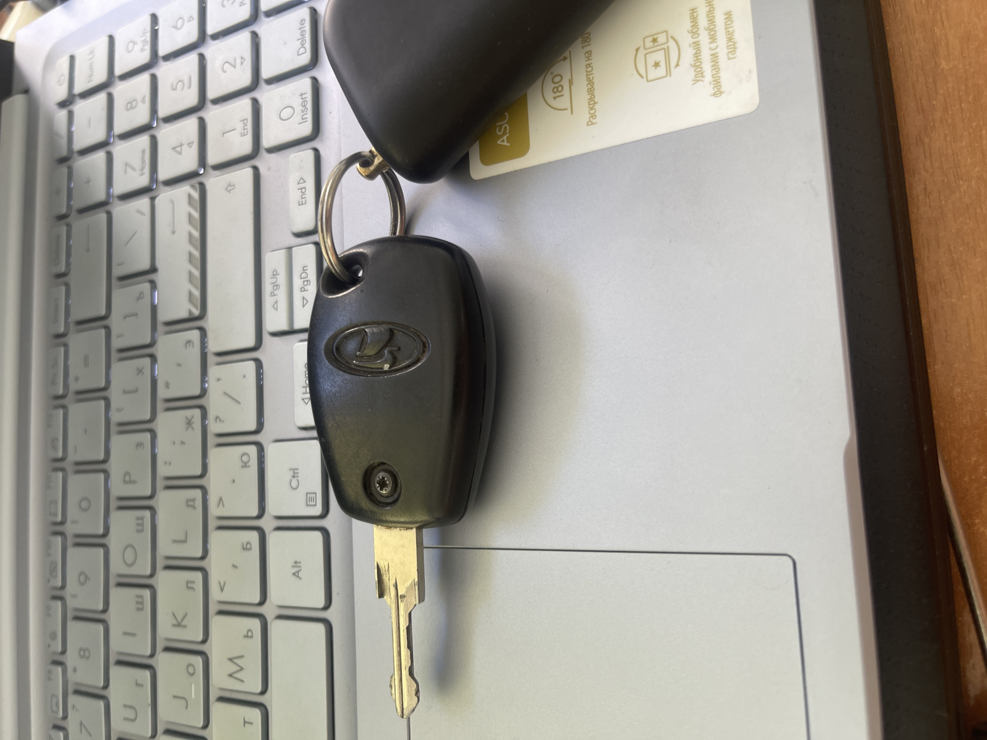 The image shows a close-up view of a car key with a Lada logo attached to a keyring, resting on a laptop keyboard. The keyboard has both Latin and Cyrillic characters visible on the keys. The laptop is a light gray color. To the right of the key, a small white card is partially visible, with Cyrillic text that appears to be instructions or promotional material. The text on the card includes "180" and "Раскрывается на 180" which translates to "Unfolds to 180". Another line reads "Удобный обмен файлами с мобильным гаджетом", meaning "Convenient file exchange with a mobile gadget". A dark object, possibly another part of the car key fob or a mouse, is also visible at the top of the frame. The scene is set indoors, likely on a desk, with a hint of a wooden surface to the right, suggesting a home or office environment. The lighting appears to be artificial, typical of an indoor setting. There are no people visible in the image. The focus is on the car key and the surrounding electronic devices, with the keyboard providing a backdrop.