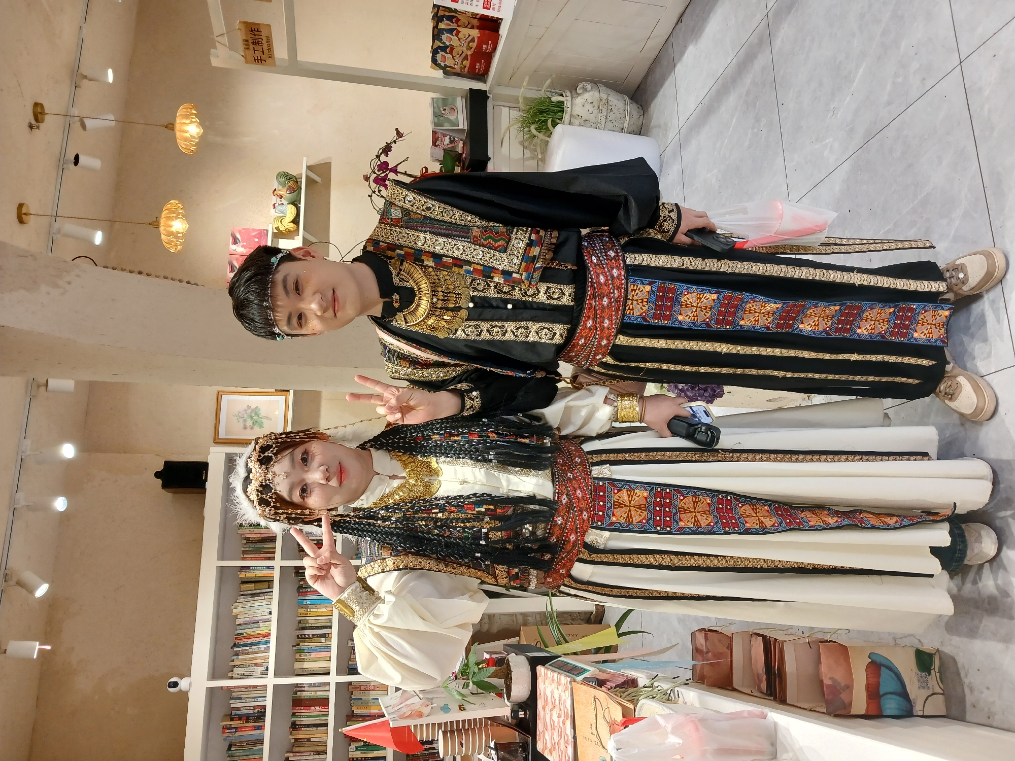 Two individuals, a female subject on the left and a male subject on the right, stand centrally posed for a photograph inside a commercial or cultural establishment in Yuzhong District, China.
The female subject wears a traditional ensemble consisting of a white long-sleeved shirt with puffed sleeves, a sleeveless tunic heavily embroidered with gold thread and multi-colored geometric patterns, a wide red and black patterned belt, and a long white skirt featuring a matching embroidered central panel. Her dark hair is styled in multiple long braids and adorned with a gold filigree headpiece incorporating white feathers. She displays multiple gold coin necklaces and gold armbands. She holds a dark-colored smartphone in her left hand and forms a 'peace' gesture with her right hand. She wears light-colored athletic shoes.
The male subject, positioned to her right, wears a complementary black and gold traditional ensemble. This includes a black long-sleeved tunic with extensive gold embroidery and multi-colored geometric patterns, a wide red and black patterned belt, and black trousers with matching vertical embroidered panels. He wears a thin light-blue headband, a prominent gold coin necklace, and gold bracelets. He forms a 'peace' gesture with his right hand and holds a clear plastic bag containing a red object in his left hand. He wears light-brown athletic shoes.
Both individuals display slight smiles towards the camera.
The indoor background features light-colored, textured walls. To the left, white bookshelves are filled with books. A small white security camera and a black speaker are mounted above the shelves. A counter in the foreground holds a small red flag, potted plants, and packaged items. To the right, overhead track lighting and two ornate amber-glass pendant lights illuminate the scene. Shelves display ceramic figures and a white display rack features a '手工制作 Handcrafted' sign in Chinese and English. The floor is covered with light grey tiles.