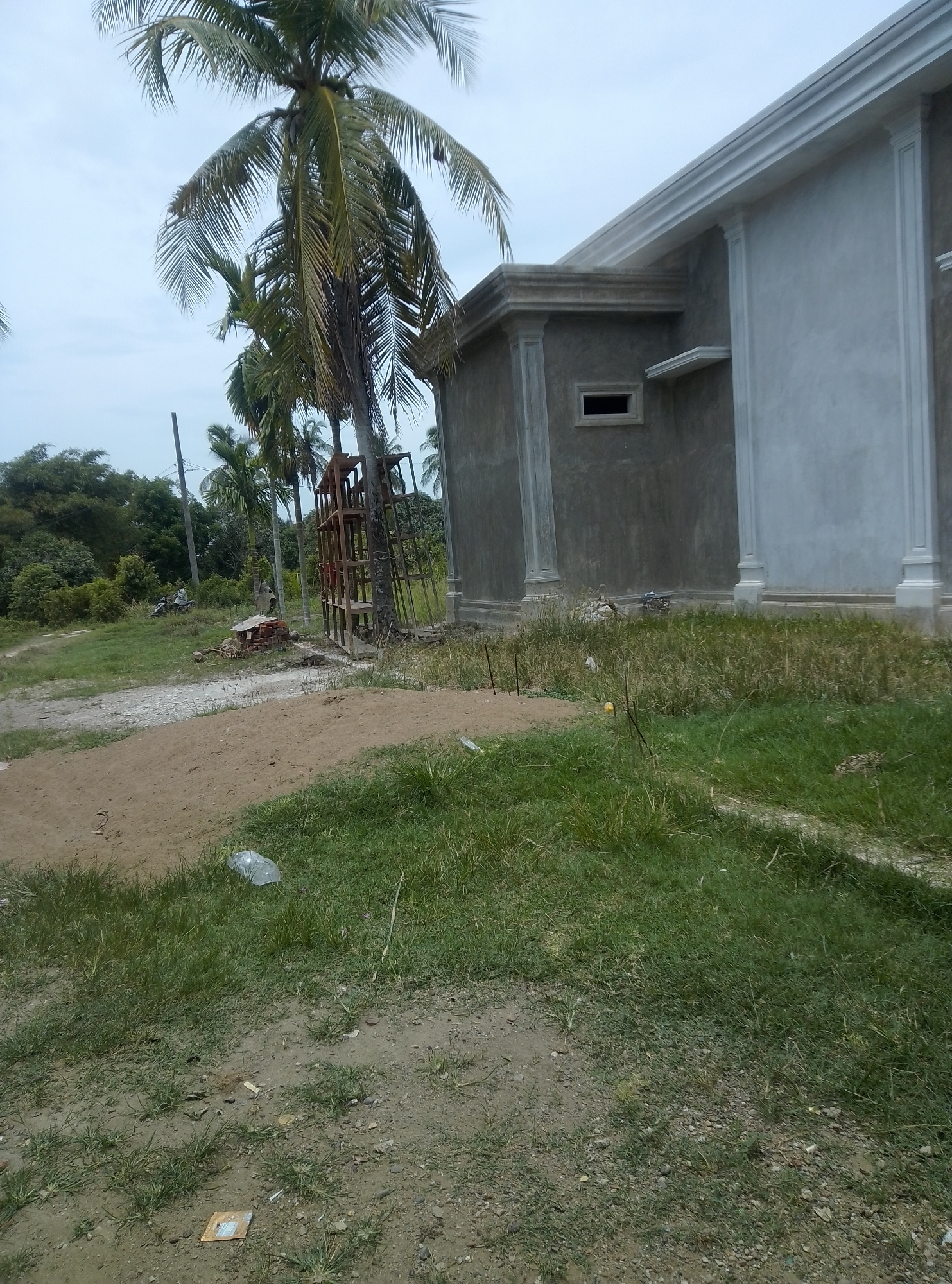 This image depicts an unfinished concrete building in an outdoor, rural setting. The building features decorative molding around the roofline and pilasters on its facade. A small, recessed window is visible on one section of the wall. In the foreground, a pile of sand and sparse green grass are present, with some debris scattered on the ground, including a plastic bag and what appears to be a piece of cardboard. Several palm trees are prominent in the scene, with one large palm tree dominating the left side of the frame. Behind the building, a wooded area with more trees is visible. To the left of the building, a metal structure resembling scaffolding or a framework stands near a palm tree. Further in the background, a dirt path leads towards a dense green landscape, and a person is seen riding a motorcycle. The sky is overcast.