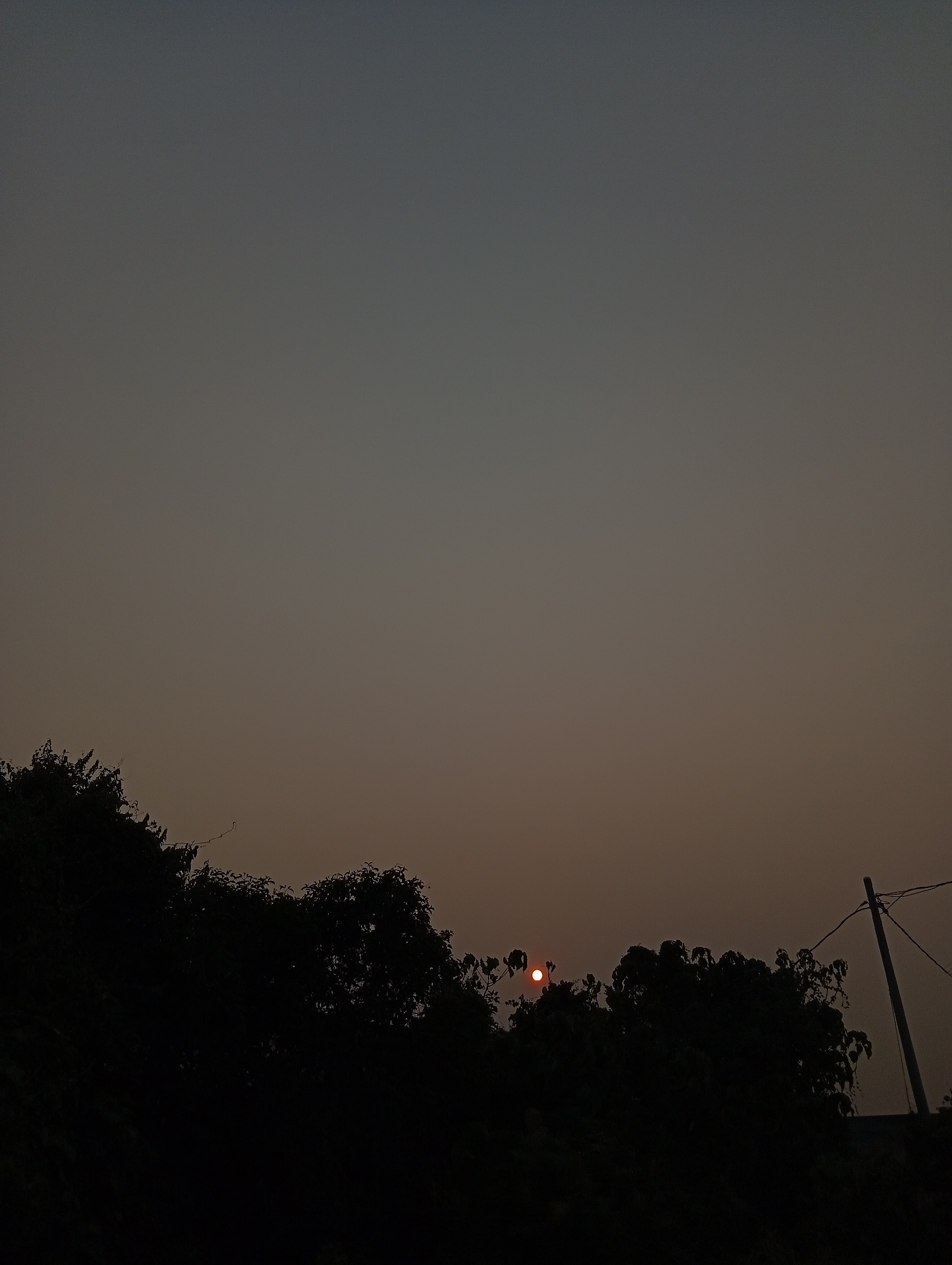 The image depicts a sunset scene with a prominent orange sun visible just above a silhouette of trees. The sky is a gradient of dark grey to lighter grey, indicating the time of day. In the lower right portion of the image, a utility pole with wires is visible, also silhouetted against the sky. The foreground is dominated by dark, shadowy trees, with some foliage discernible. The overall mood of the image is calm and serene. The image is believed to have been taken in the city of Kualu, Indonesia, although no specific landmarks are visible to confirm this location.