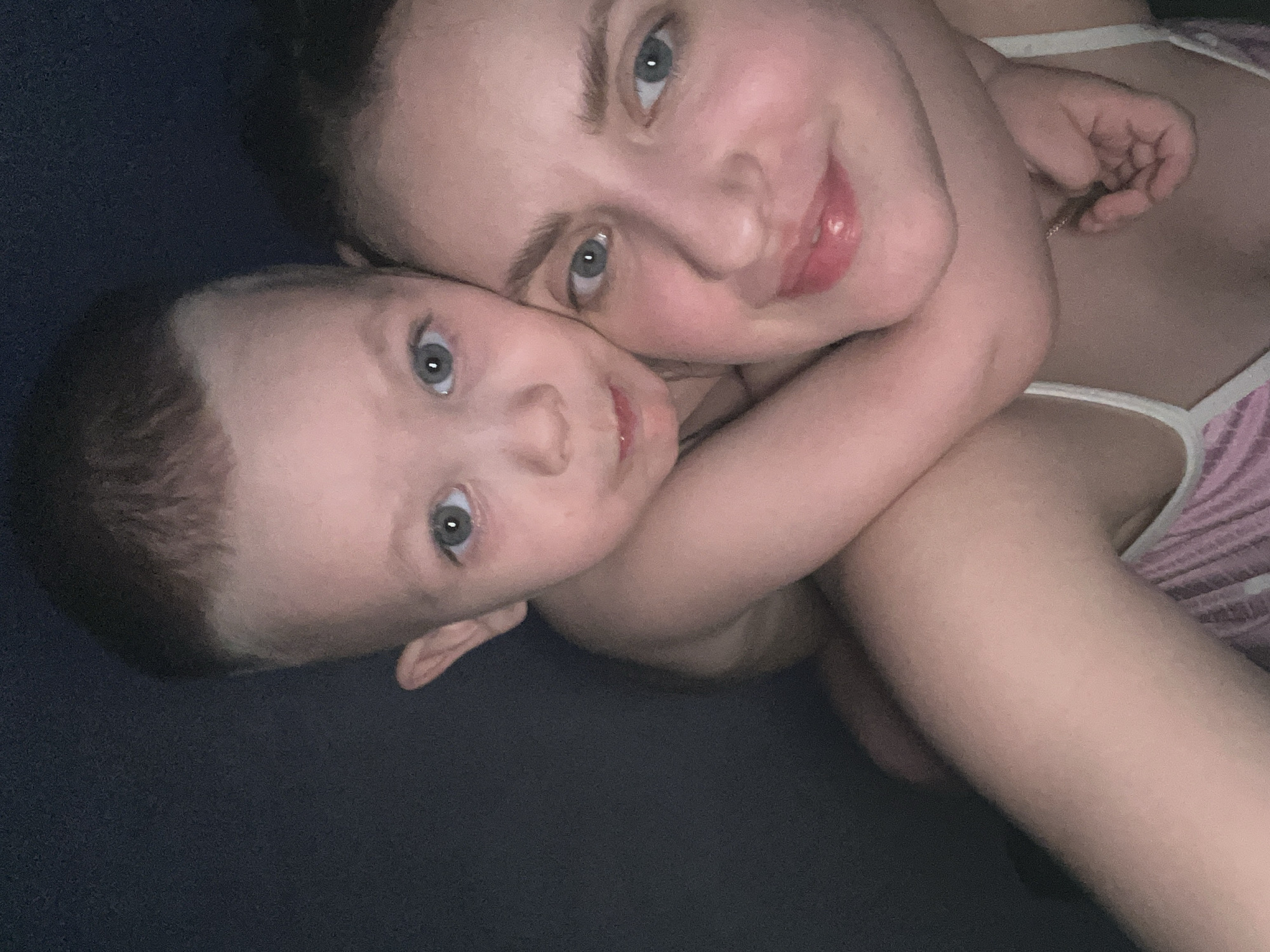 This intimate, close-up portrait features an adult woman and a young child, likely a mother and her baby, lying together in a tender embrace. Both individuals have fair skin and captivating blue-gray eyes, looking directly towards the camera.
The woman is positioned slightly behind and to the right, exhibiting a gentle smile and dark hair that appears pulled back. She is wearing a light-colored, possibly white or pale pink, strap top with subtle vertical stripes visible on the right side of the frame.
The young child is in the foreground, positioned to the left. The child has light brown hair and a soft, inquiring expression. Their left arm is affectionately wrapped around the woman's neck or shoulder, emphasizing their close bond.
The setting appears to be indoors, against a solid dark blue or black background, likely a bed or sofa, which creates a cozy and intimate atmosphere. The lighting is soft and dim, suggesting either low ambient light or artificial light, enhancing the warmth of the scene.
The image powerfully conveys emotions of love, comfort, and deep familial connection. There are no specific external location cues visible within the image itself that would identify the context as Марковский, Russia. No text is visible in the media file.