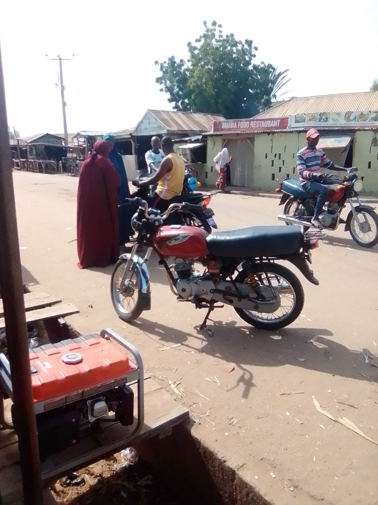 A group of people are gathered outside a building in Talasse, Nigeria. A man in a red and blue striped shirt and a red cap is sitting on a motorcycle, facing left. Behind him, another motorcycle is parked. In the background, the building has a sign that reads "AMANA FOOD RESTAURANT". To the left of the restaurant, there are market stalls. Two women in traditional Islamic clothing are standing in front of the restaurant. A portable generator with an orange top is visible in the foreground, placed on wooden planks. The ground is dusty and appears to be unpaved.